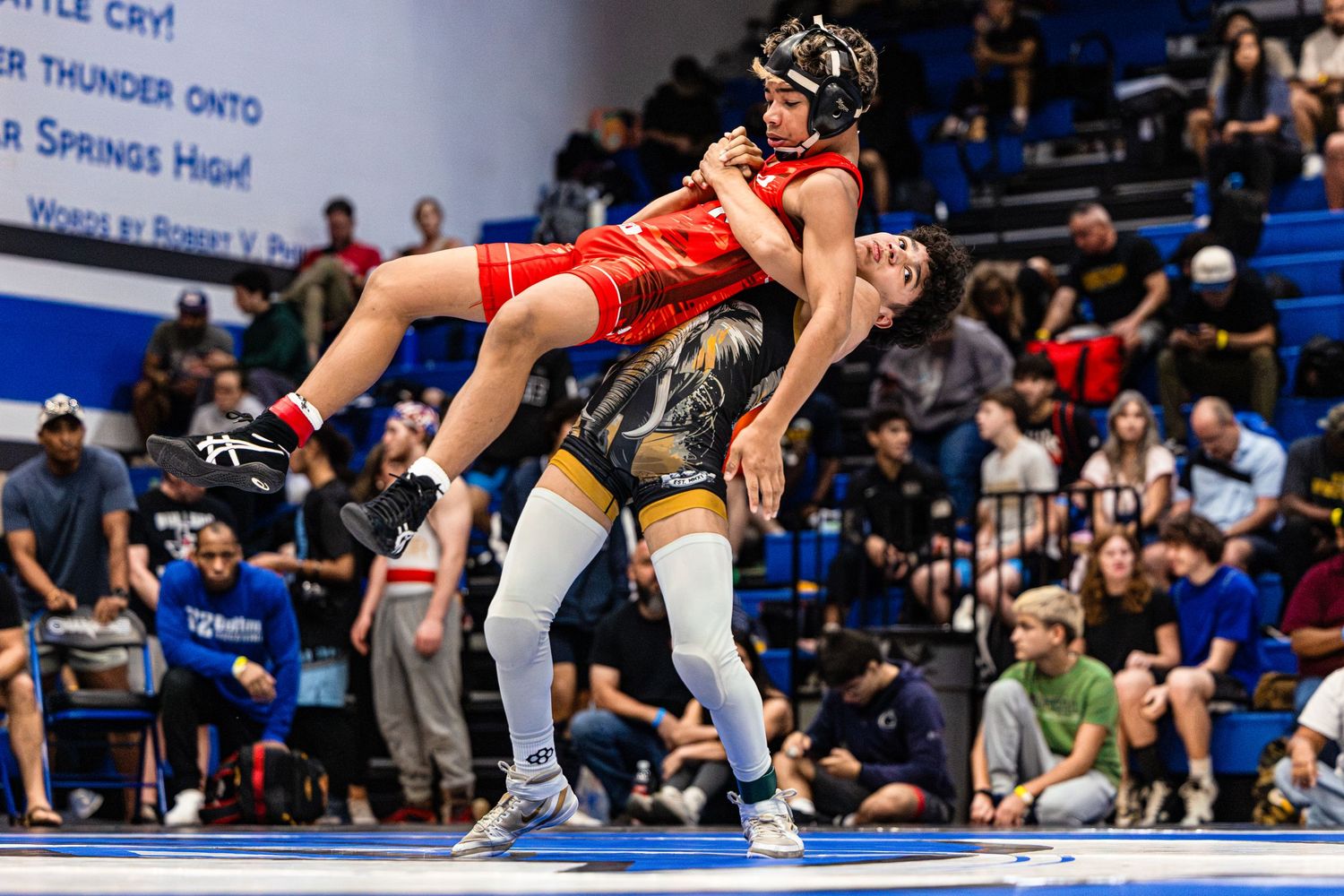 Two wrestlers compete in a high school wrestling match, one lifting opponent off the mat in a crowded gymnasium.