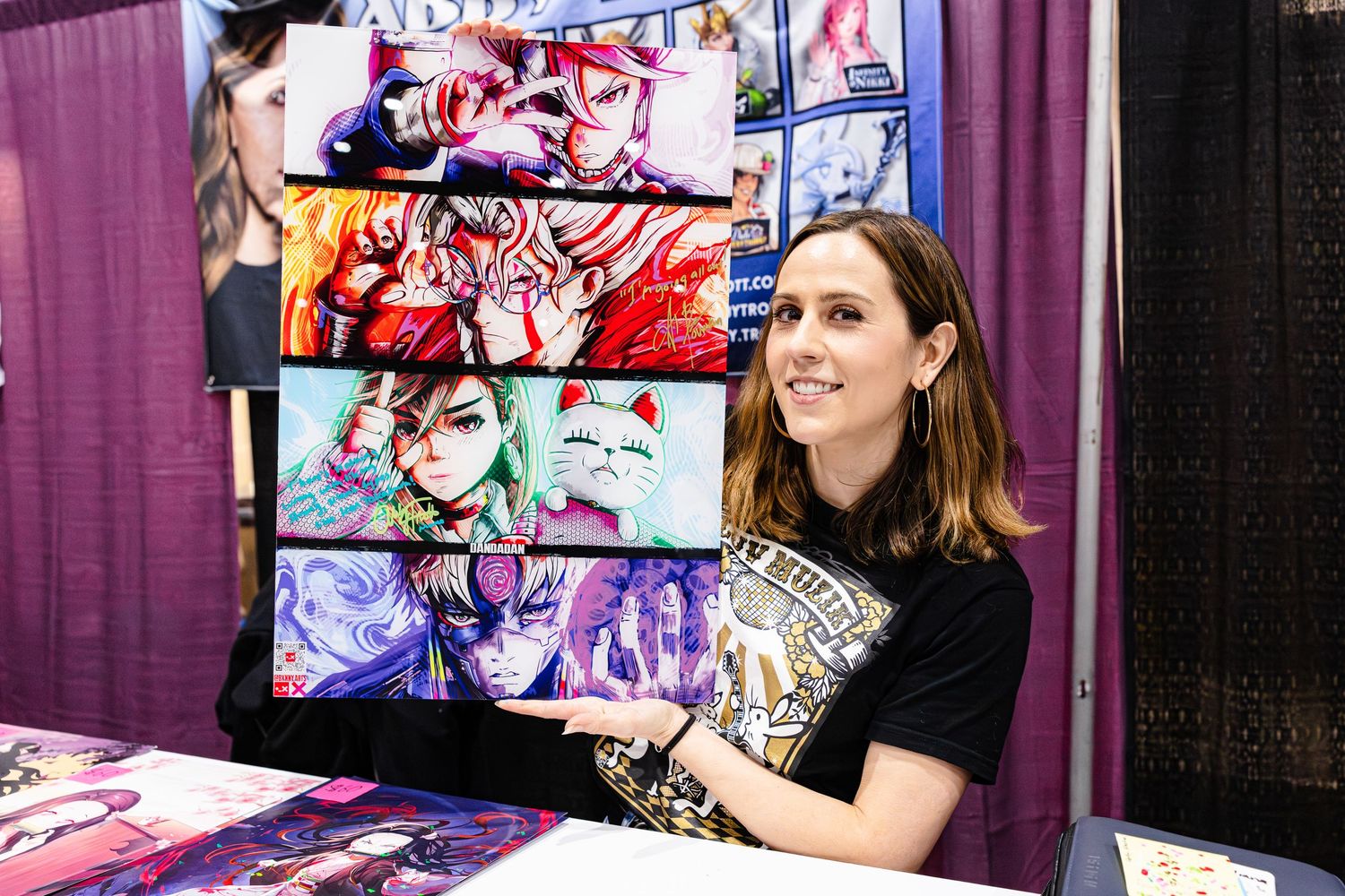 Artist proudly displays colorful anime-style artwork prints at a convention booth while seated at a table.