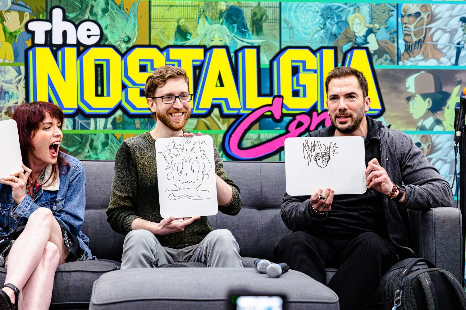 Four people on a couch holding drawings at The Nostalgia Con event, smiling and laughing together.