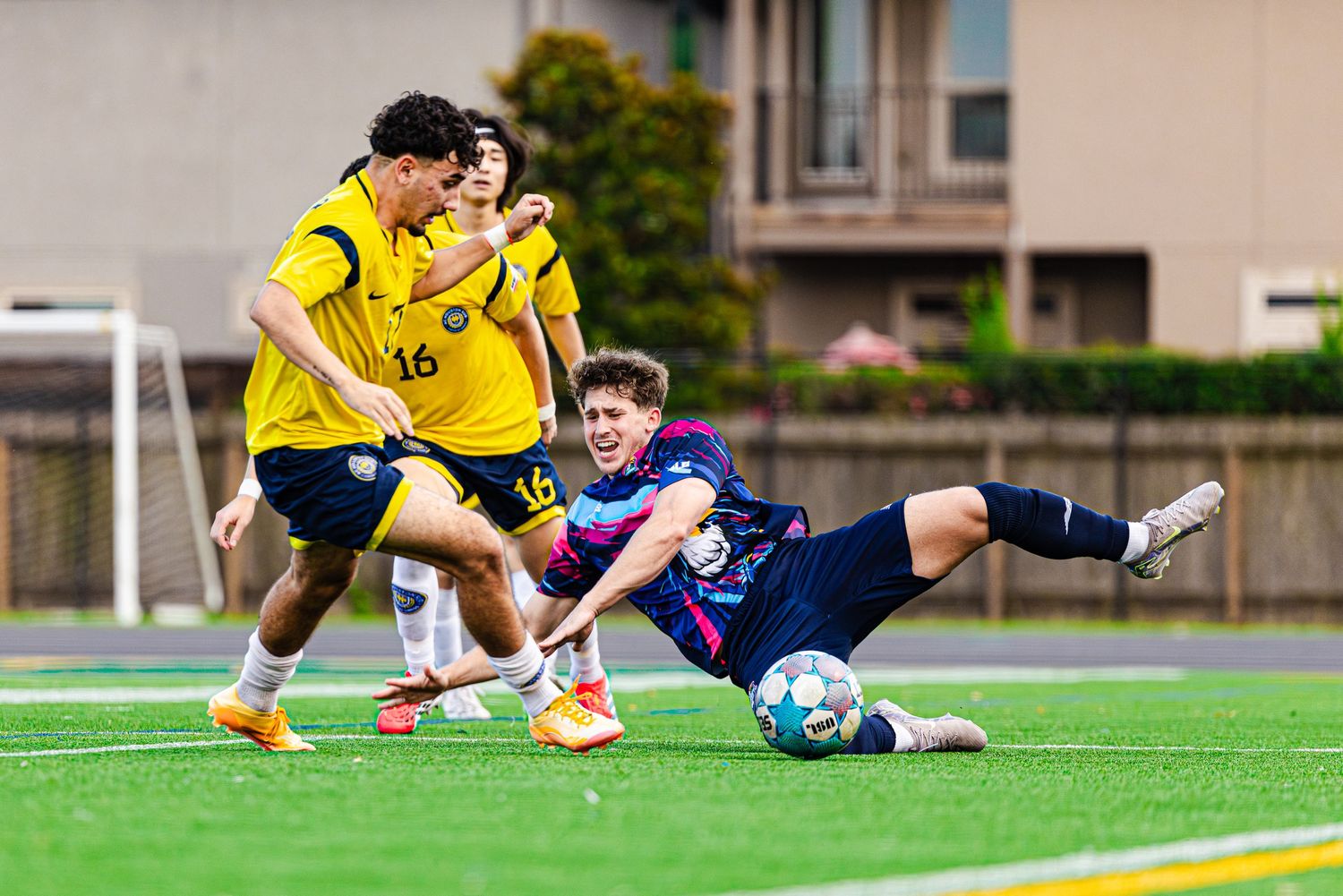 Soccer player in blue slides to challenge two opponents in yellow jerseys during a competitive match on a green turf field.
