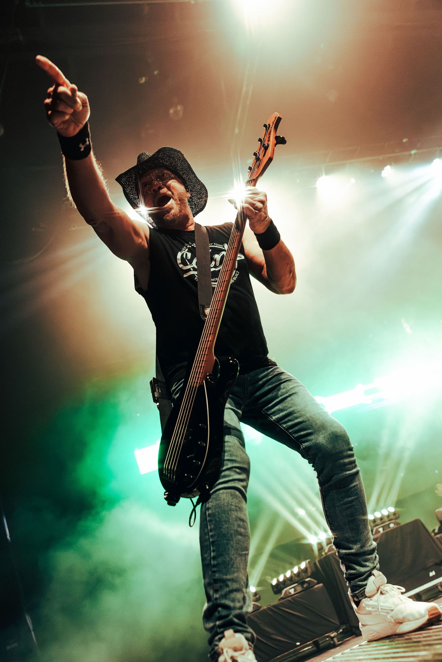 A rock guitarist performs energetically on stage, wearing a black hat and sleeveless shirt, with green and orange stage lights behind him.