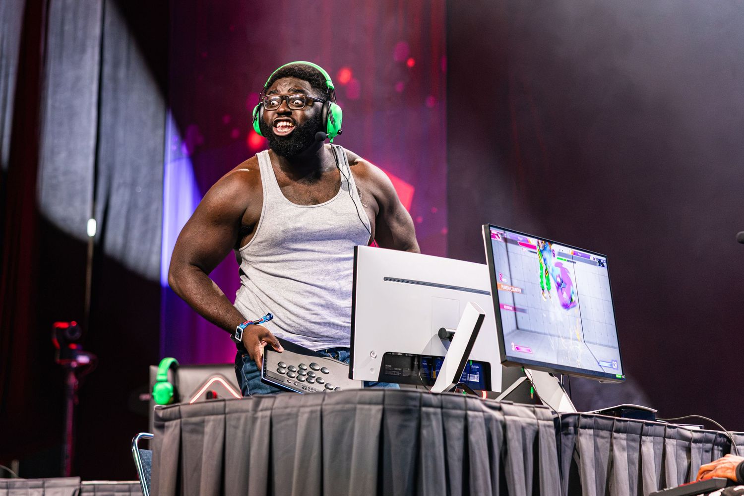 A smiling DJ wearing green headphones performs at a music event behind a DJ booth with monitors.
