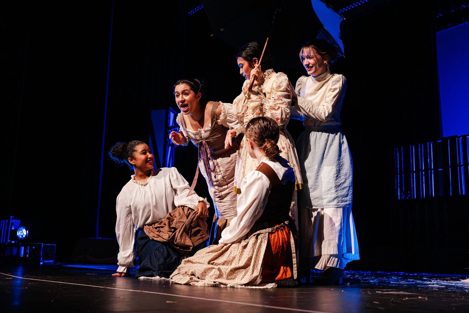 Theater performers in period costumes on stage during a dramatic scene under blue and white lighting.