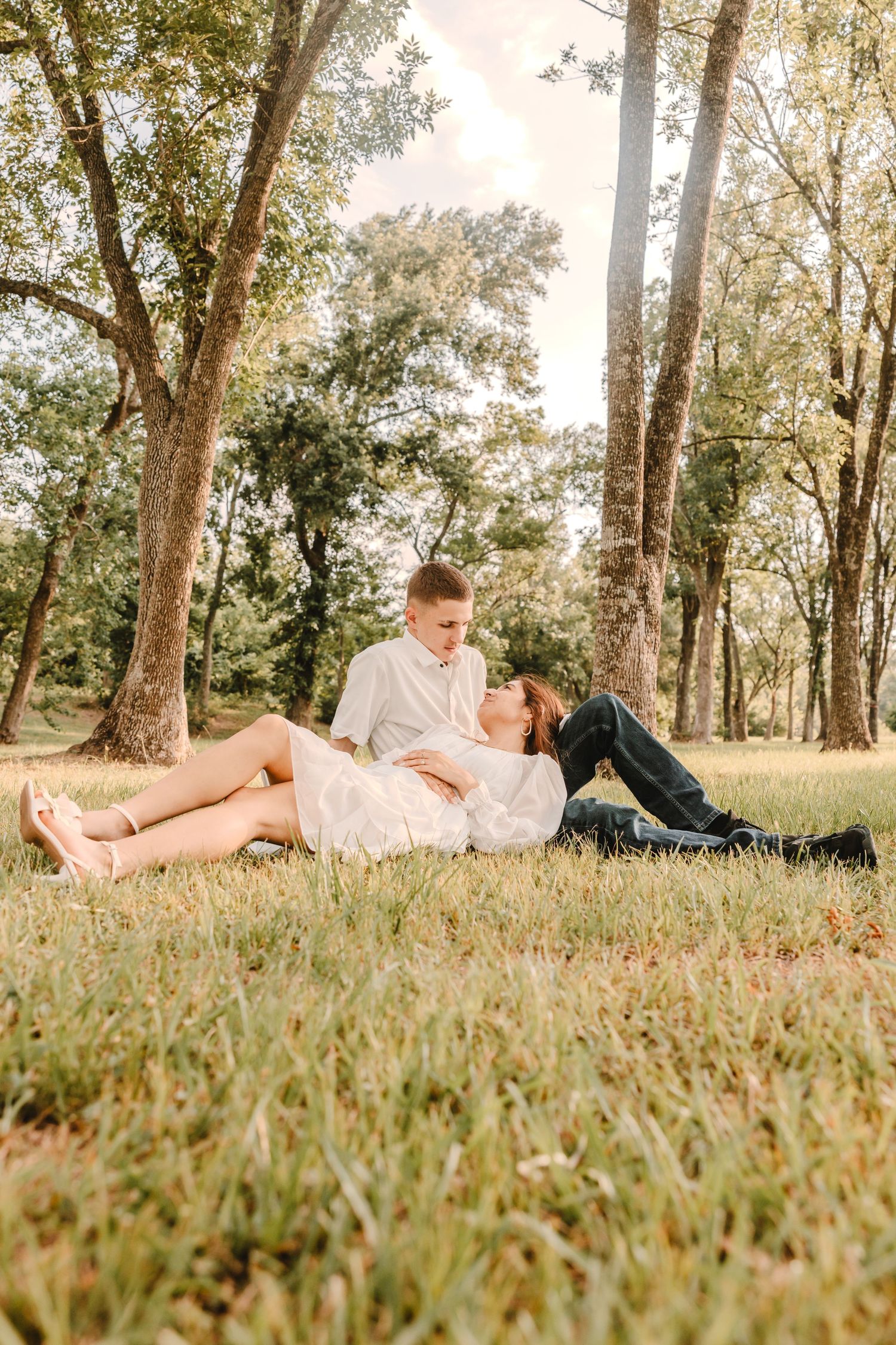 A couple lounges on grass during a romantic golden hour photoshoot among trees.