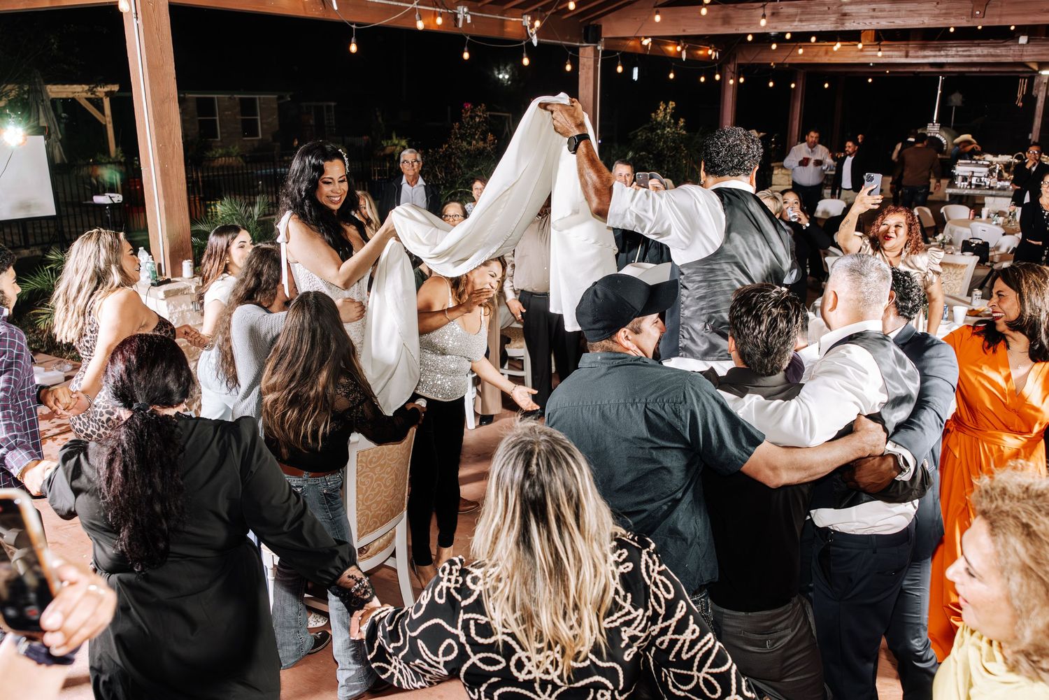 Wedding guests dance and celebrate together on a lively dance floor under twinkling lights at an indoor reception.