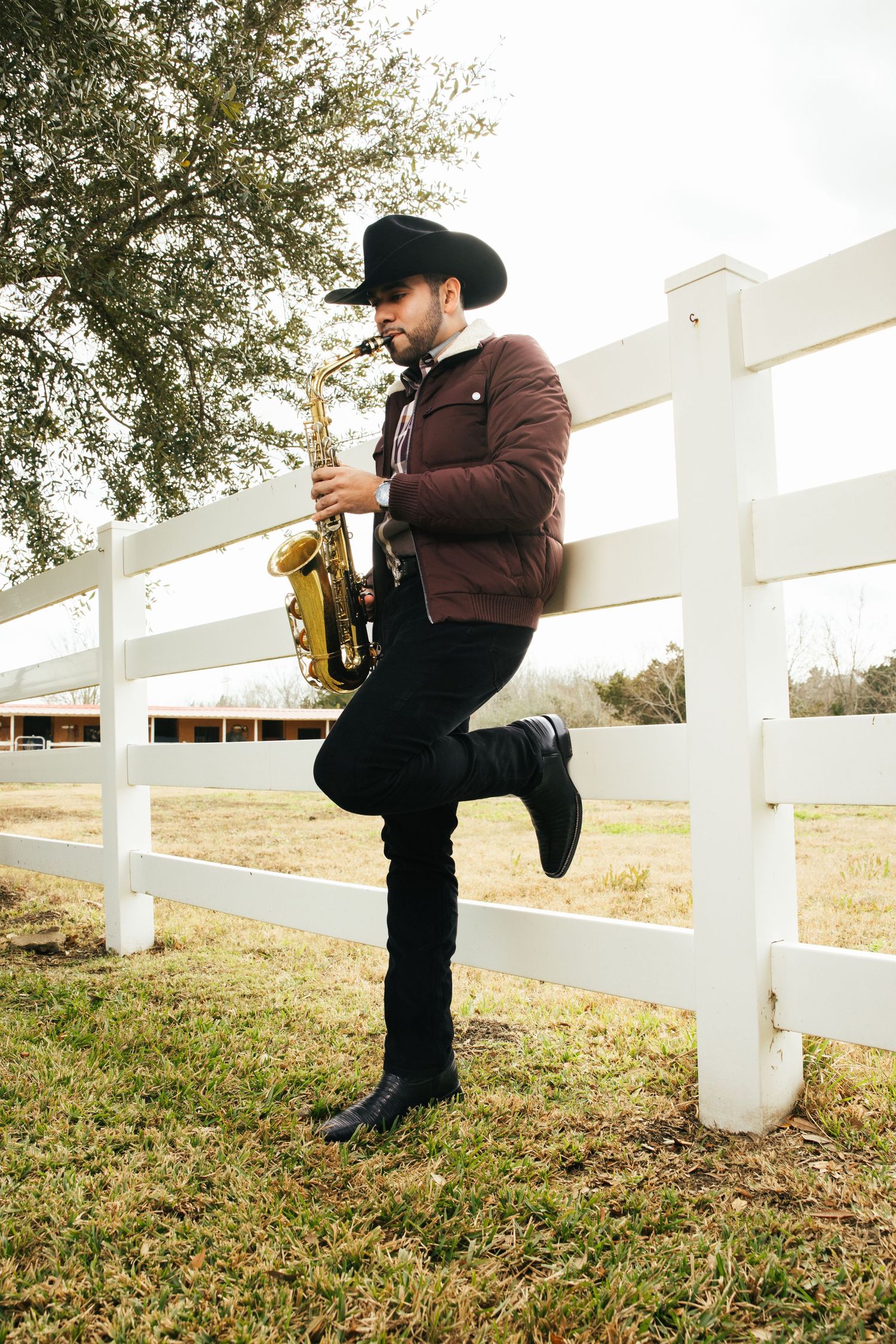 Musician in brown jacket and cowboy hat plays saxophone while leaning against white fence in countryside setting.