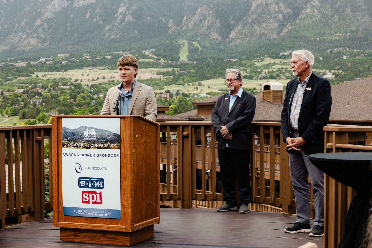 Young man speaking at outdoor podium with two men standing behind him, mountains and trees visible in background.