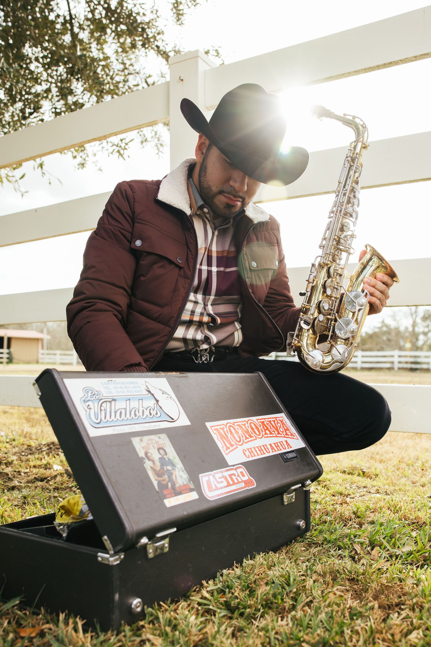 A musician in western attire sits on grass with a saxophone and open case displaying stickers in sunlit outdoor setting.