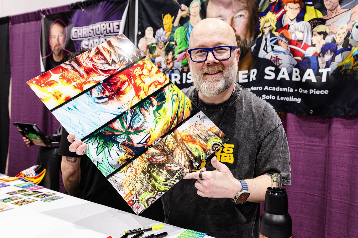 Christopher Sabat smiles holding anime artwork prints at his signing booth at a fan convention.