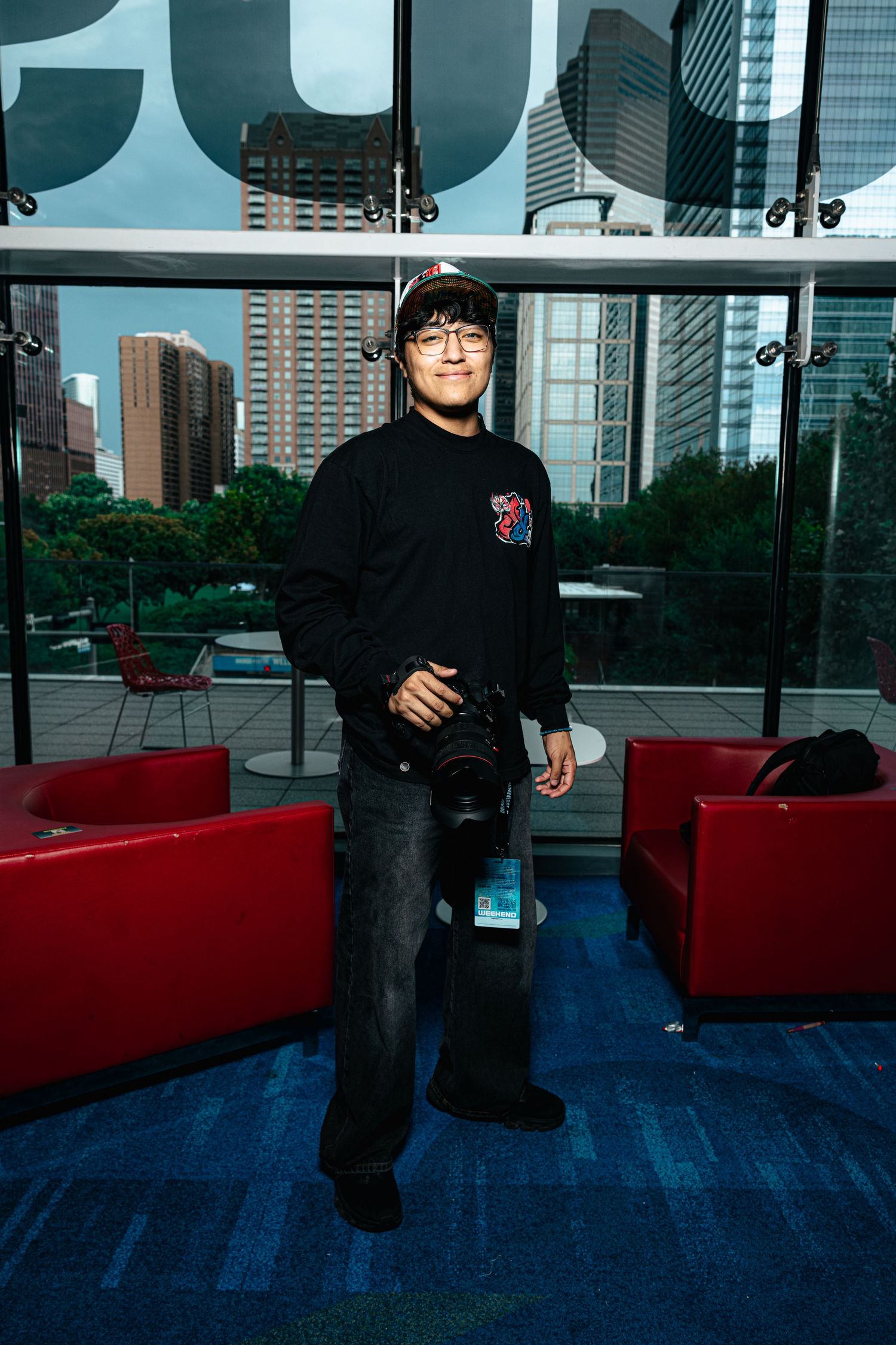Young photographer holding camera indoors, wearing black sweatshirt and cap, city skyline visible through windows behind him.