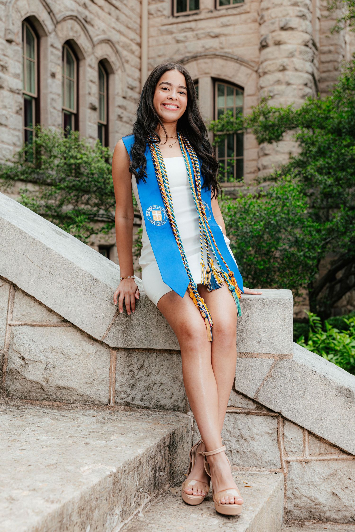 Smiling graduate poses on stone steps wearing blue sash and honor cords outside historic university building.
