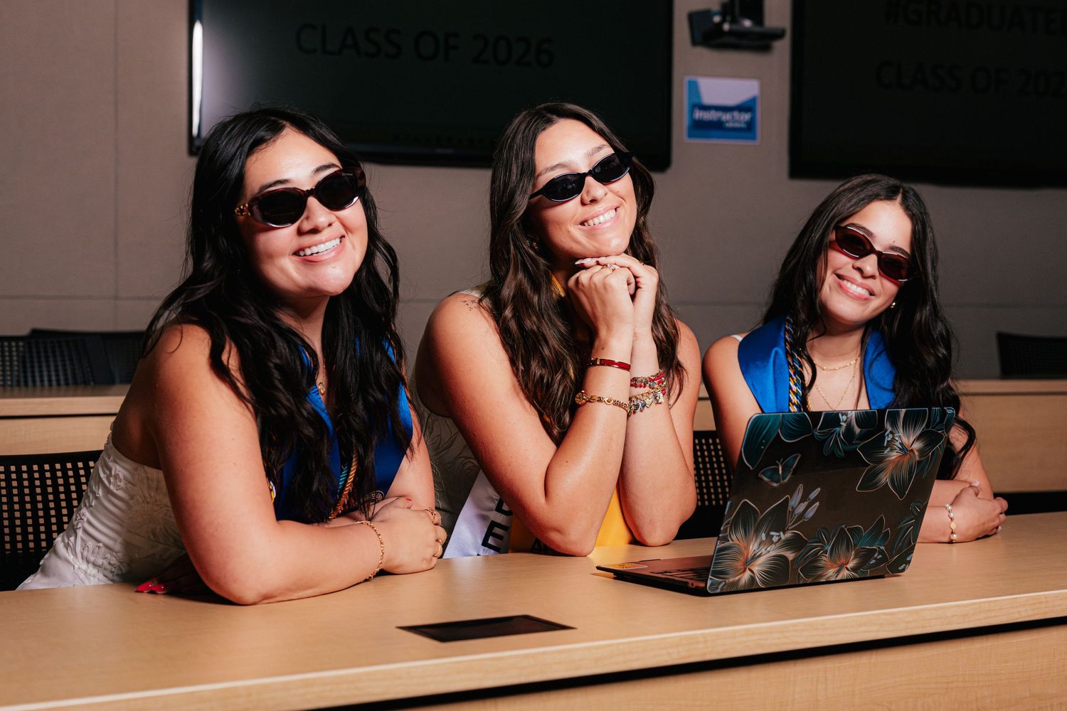 Three smiling young women wearing sunglasses sit together at a classroom desk with a laptop, class of 2025 sign behind them.