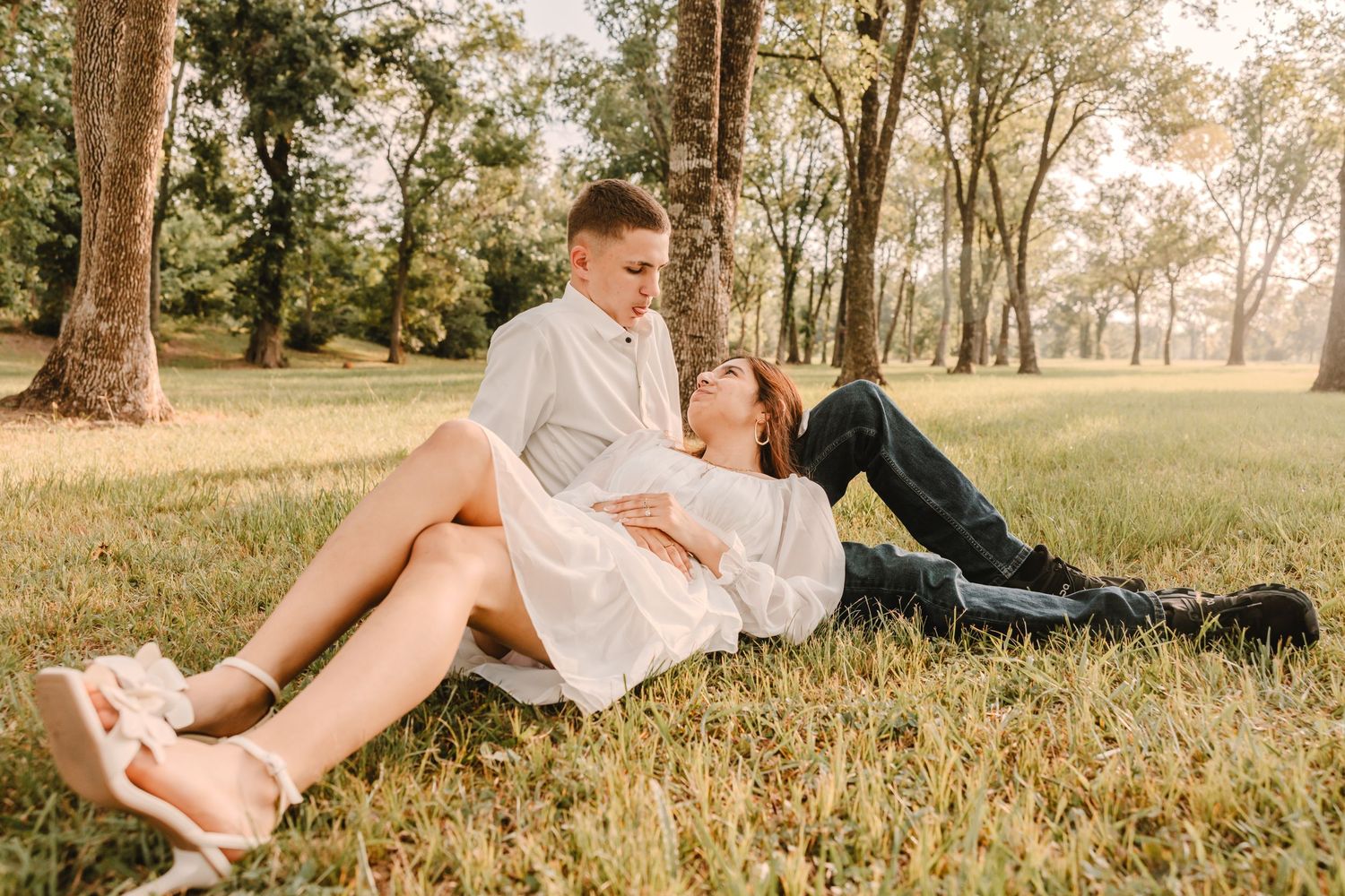 A couple lounges together on a grassy hillside during a romantic outdoor photoshoot at sunset.