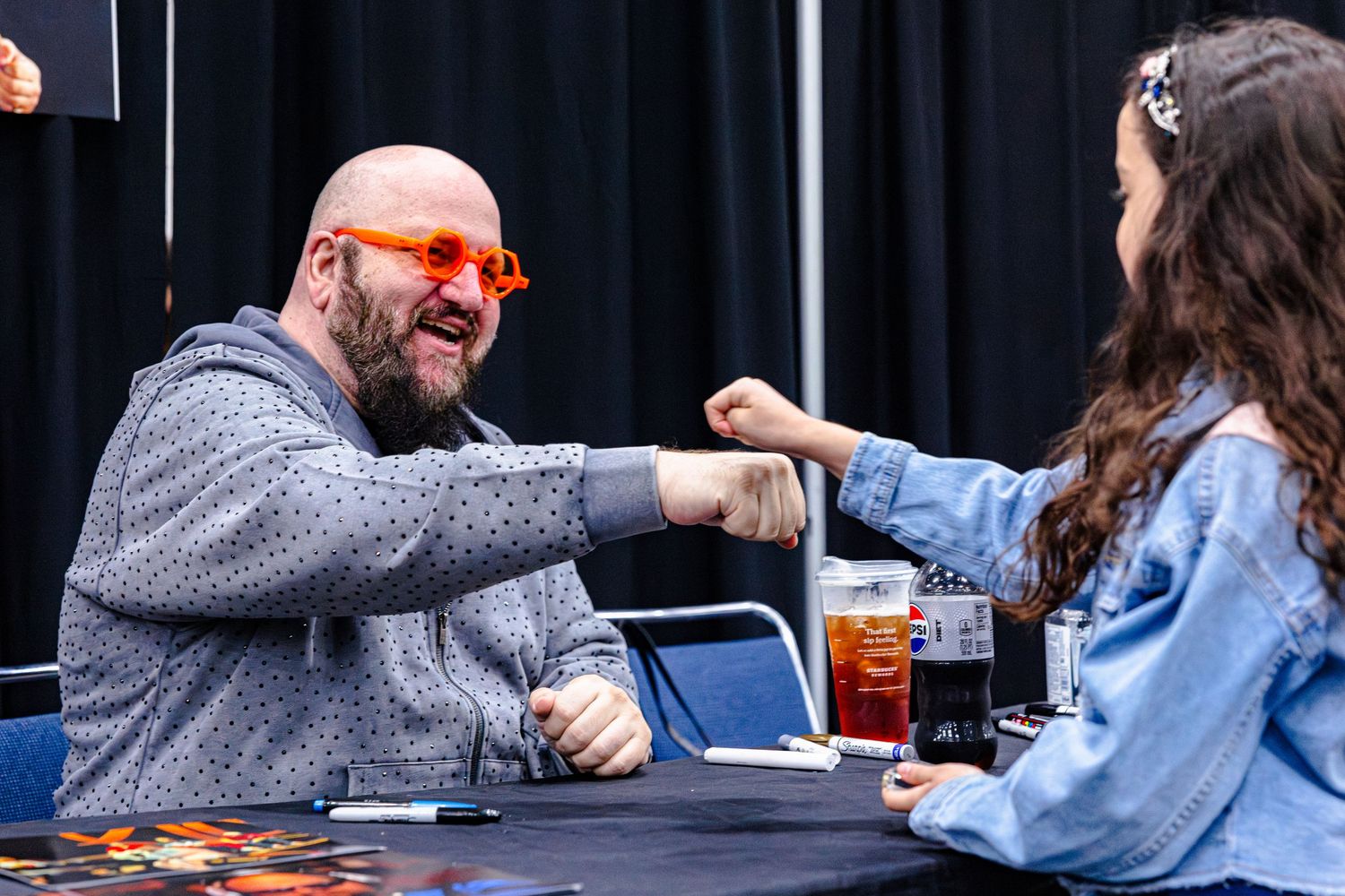 Bald man with orange glasses fist-bumping a young woman at a convention signing table.