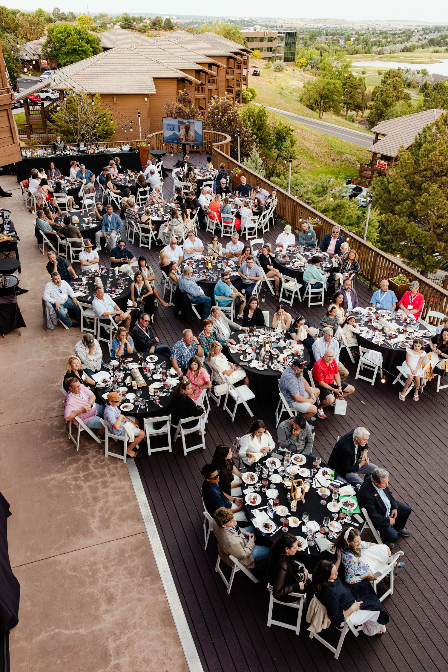 Aerial view of elegant outdoor wedding reception on a deck with round tables and white chairs surrounded by trees.