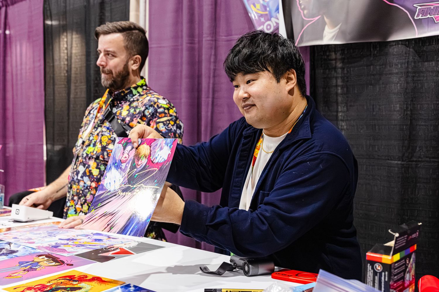 Two men smile while displaying vibrant anime artwork prints at a convention booth.