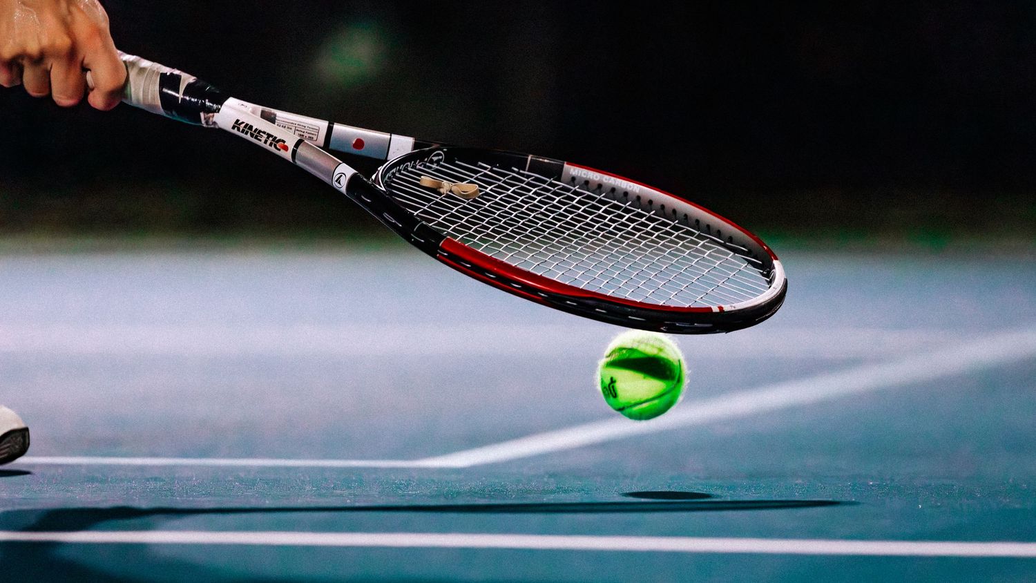 Close-up shot of a tennis racket hitting a green tennis ball on a blue court surface.