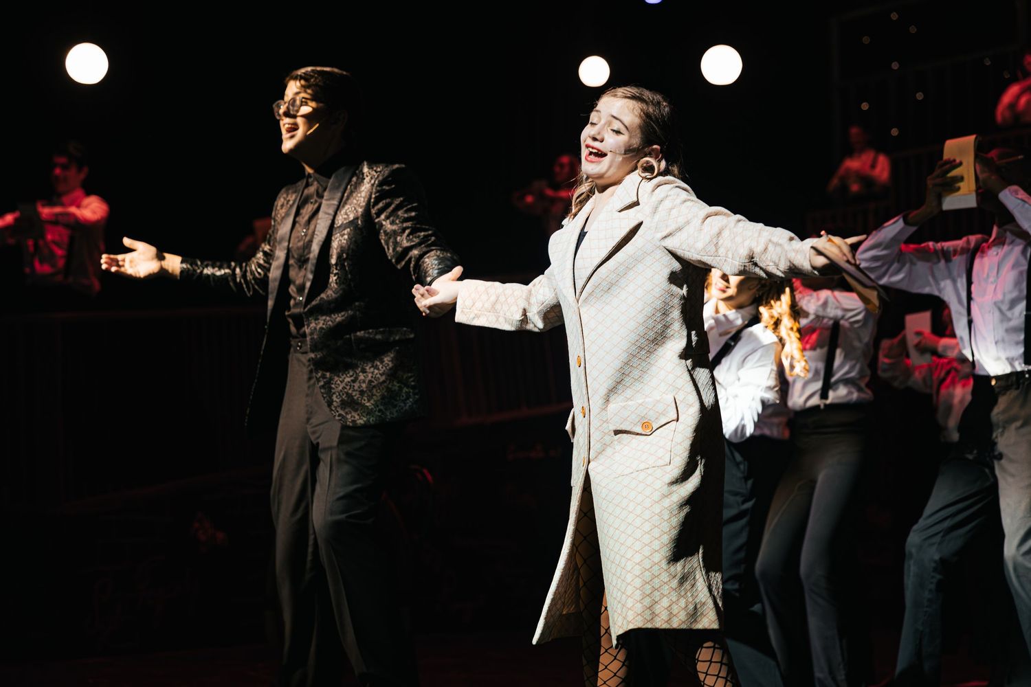 Performers in white and black outfits dance and gesture dramatically on a dark stage during a theatrical performance.