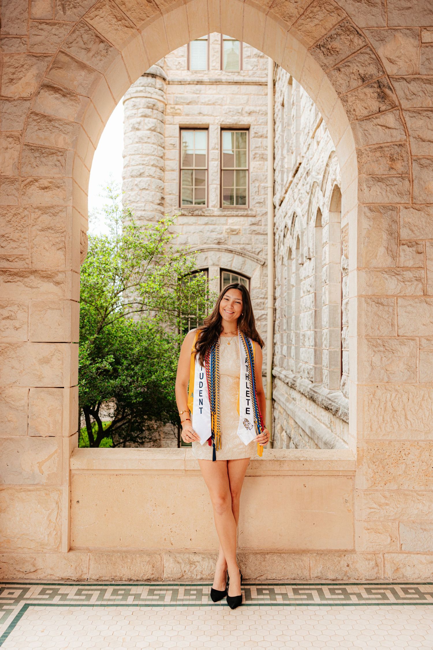 Graduate in white dress and academic stoles poses under stone arch at university building.
