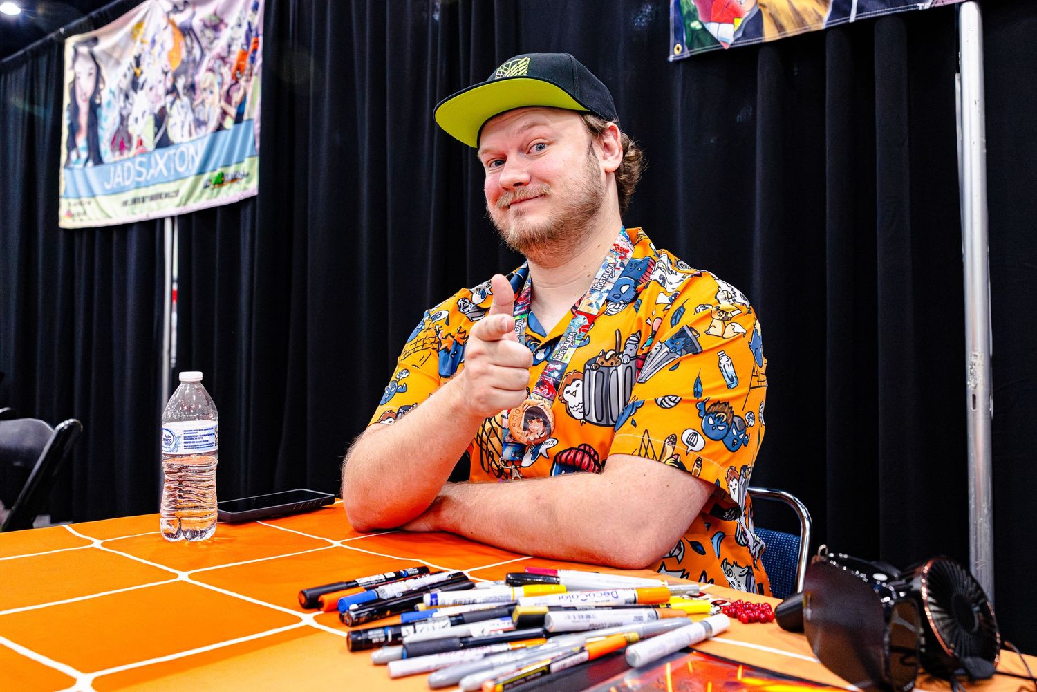 Man in colorful Hawaiian shirt and green cap sits at signing table with markers at a comic convention.