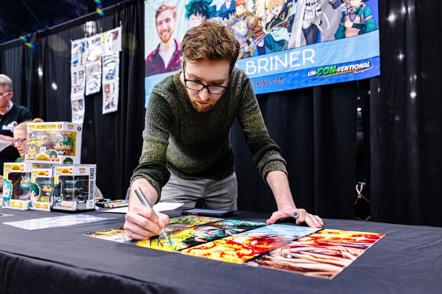Artist signing a large colorful print at a comic convention booth with collectibles displayed.
