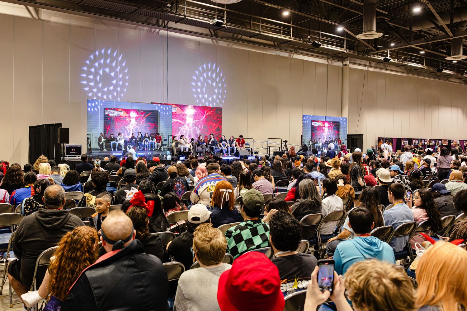 Large crowd gathered at an indoor convention hall watching a live stage performance with colorful lighting.