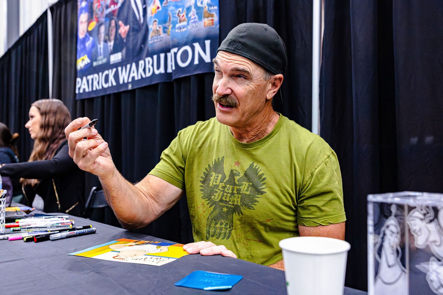 Patrick Warburton signing autographs at a convention table, wearing a green t-shirt and black beanie.