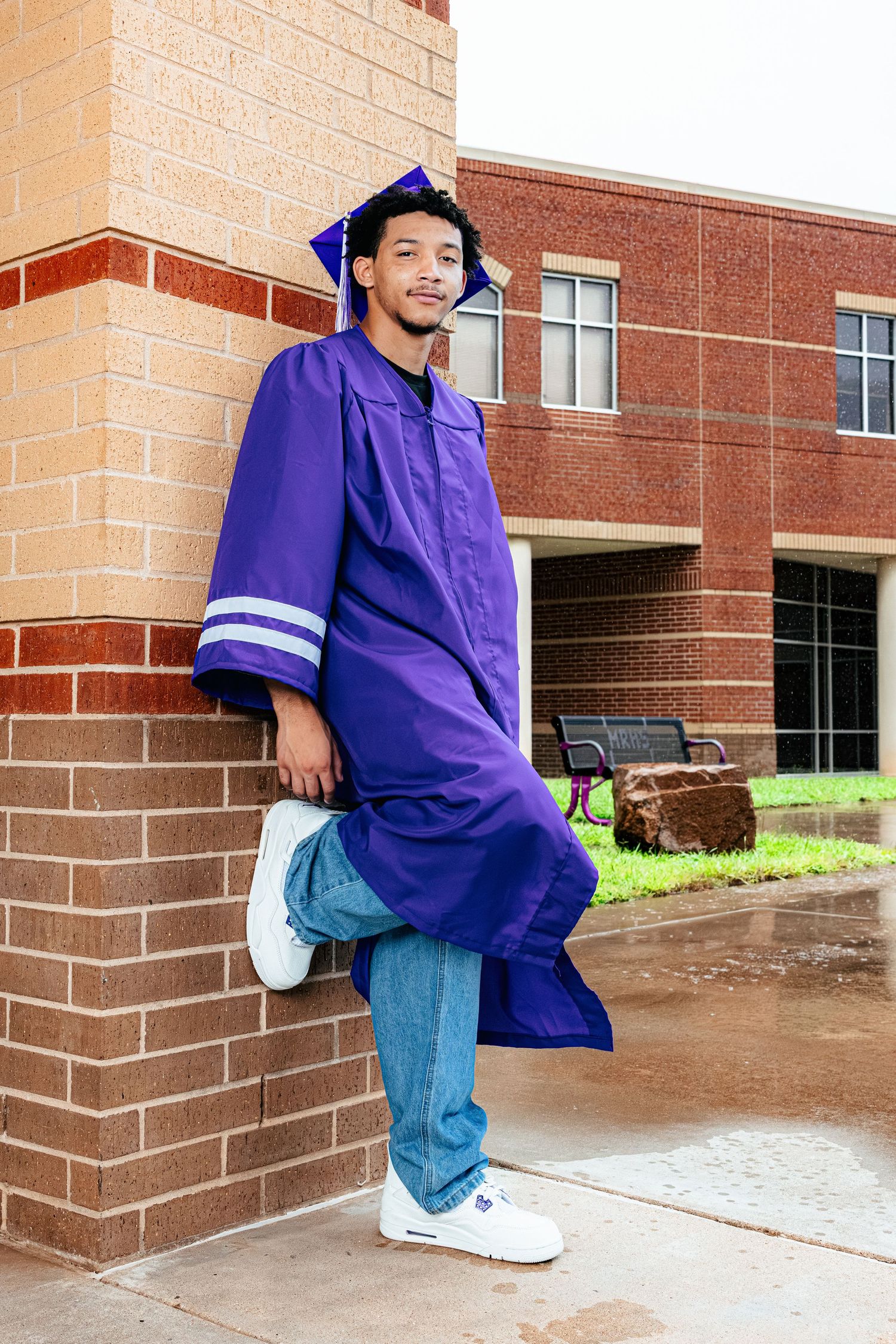 Young man in purple graduation cap and gown leaning against brick wall outside school building, smiling proudly.