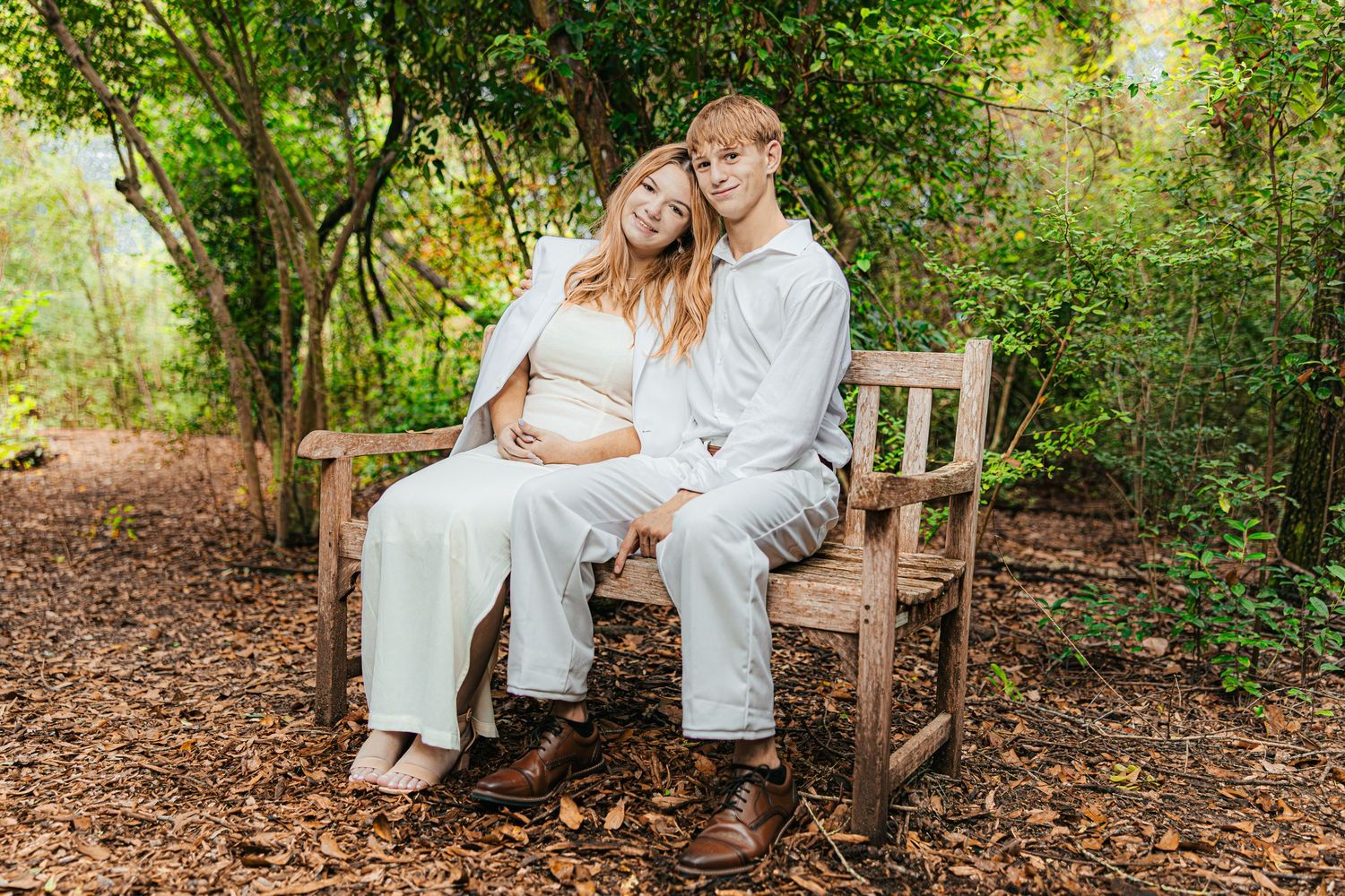 A couple shares a tender moment while sitting together on a rustic wooden bench in a wooded garden setting.