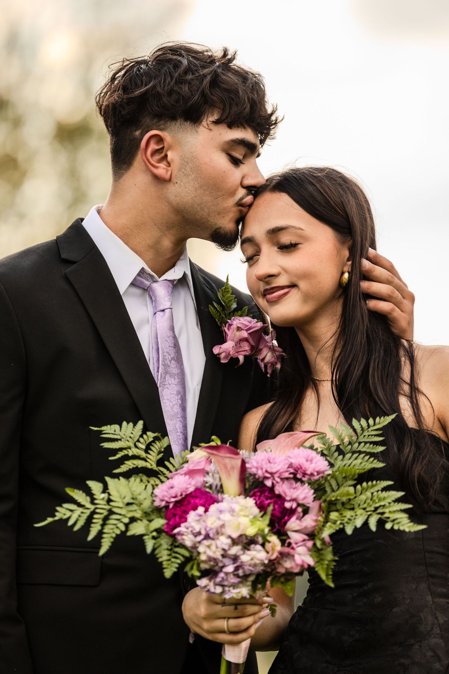 Romantic engagement photo with couple holding pink flowers and ferns in formal attire.