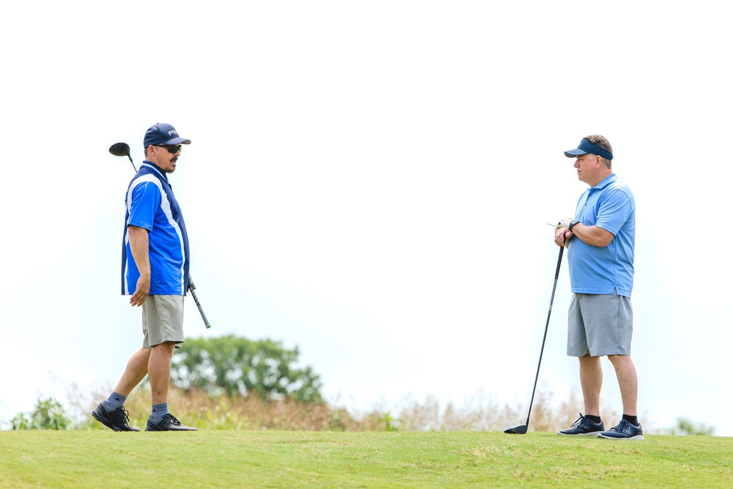 Two golfers in blue shirts and shorts stand on a grassy hill with golf clubs during a sunny day.