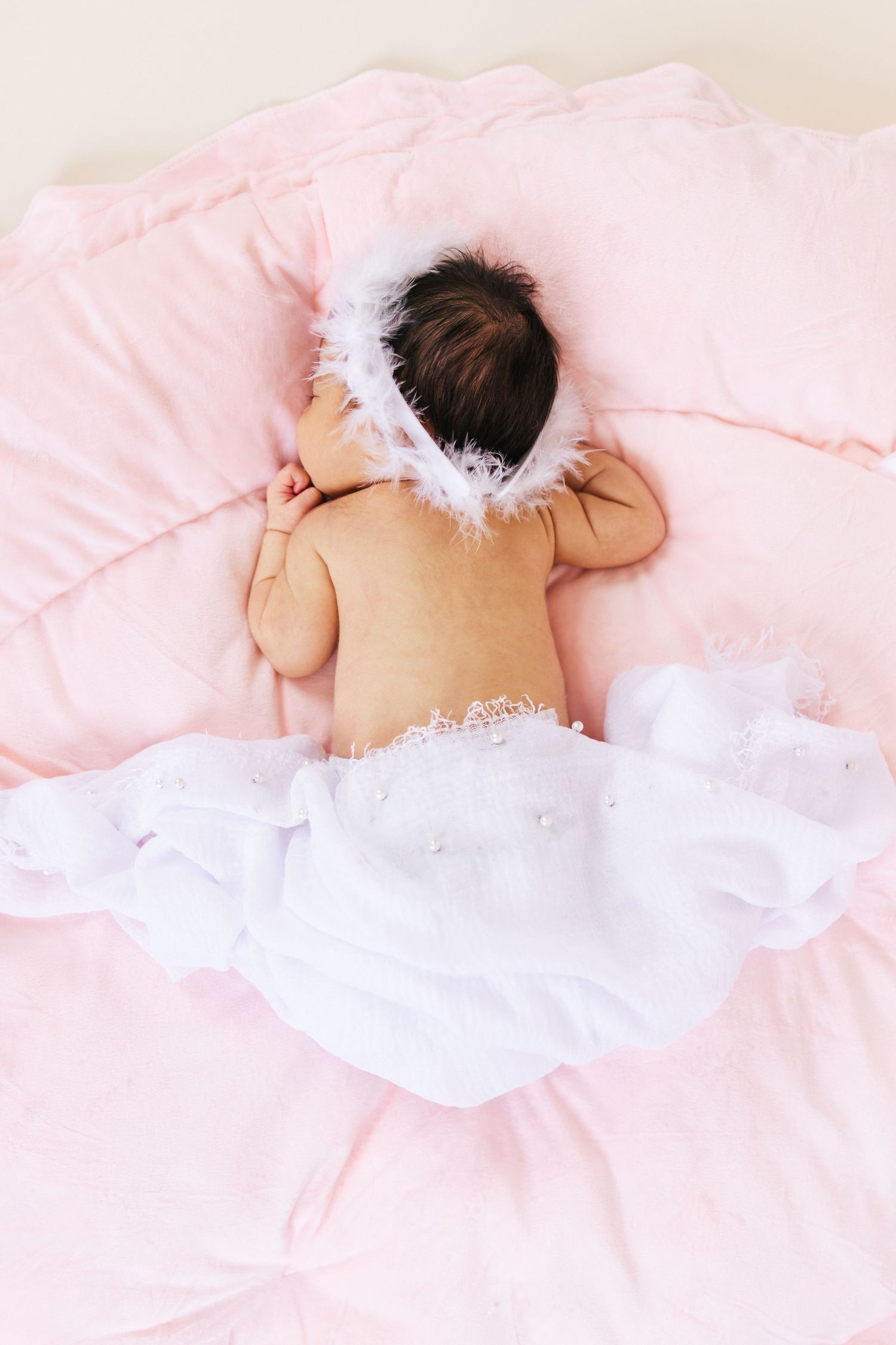 A sleeping baby wearing angel wings lies on soft pink bedding in a dreamy nursery setting.