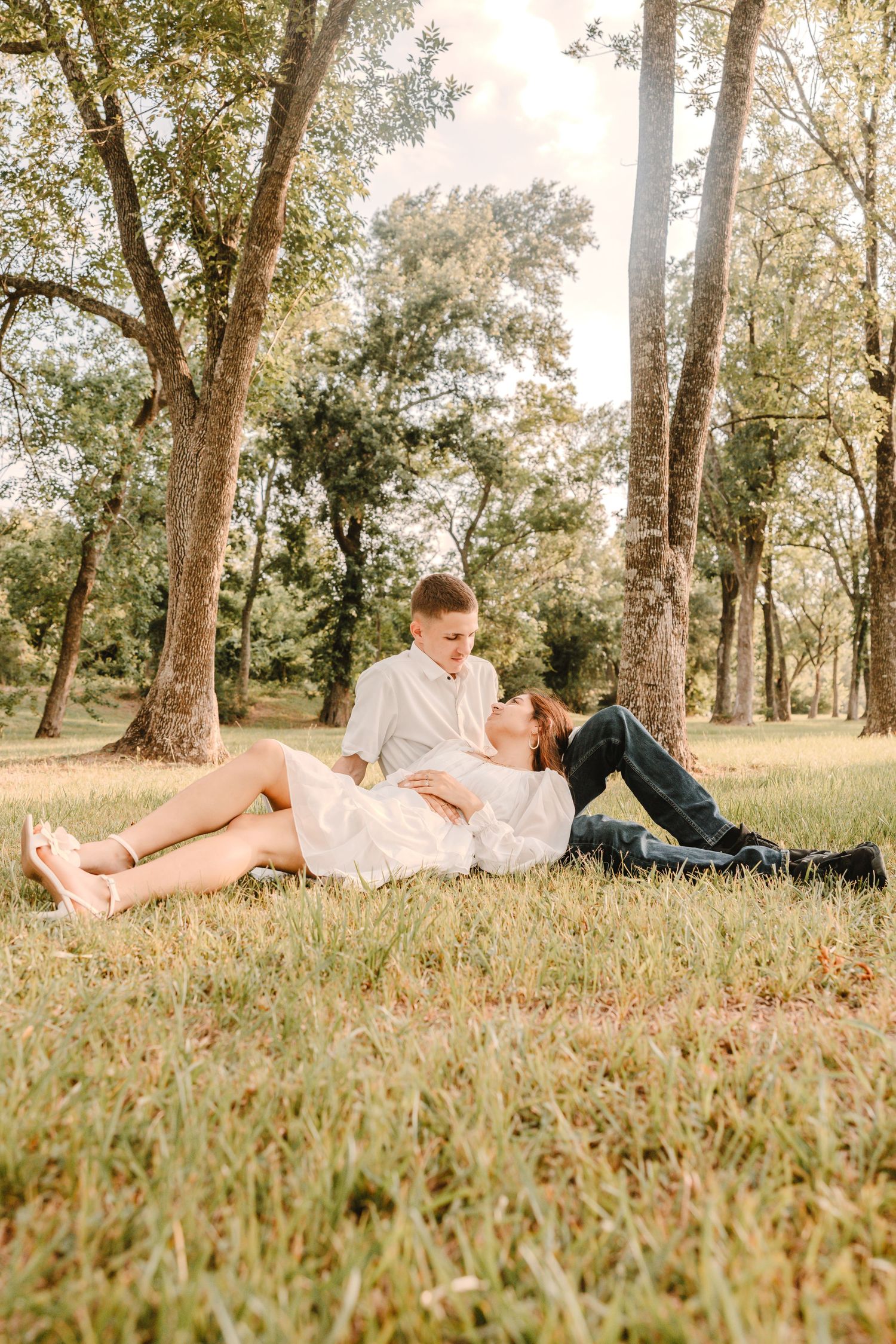 Romantic couple lying in grass under tall trees in a park during golden hour sunlight.