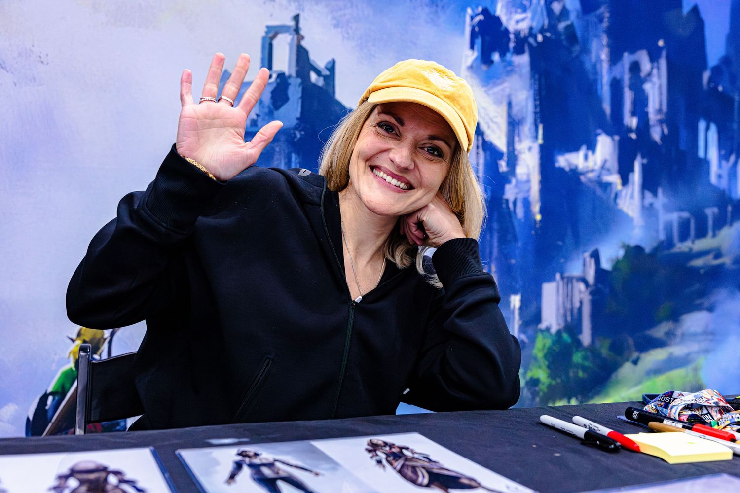 Smiling blonde woman in yellow cap waves at camera while seated at convention signing table.
