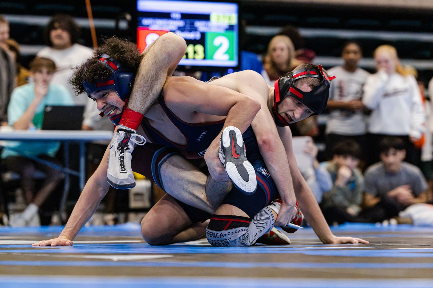 Two wrestlers compete on a blue mat during a high school wrestling tournament match.