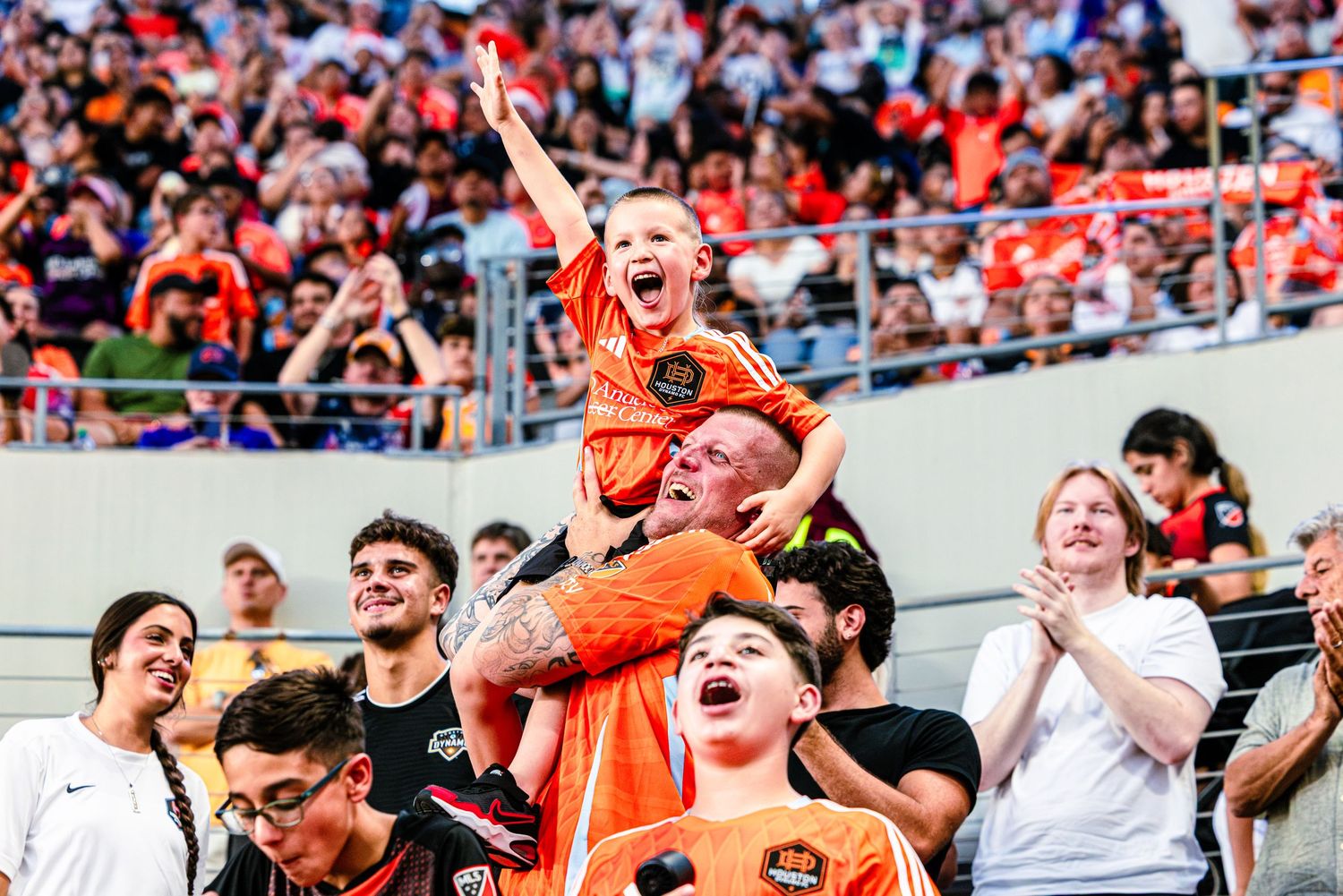 Excited Brisbane Roar fans in orange jerseys celebrate at a packed soccer stadium, with a child raised on shoulders.