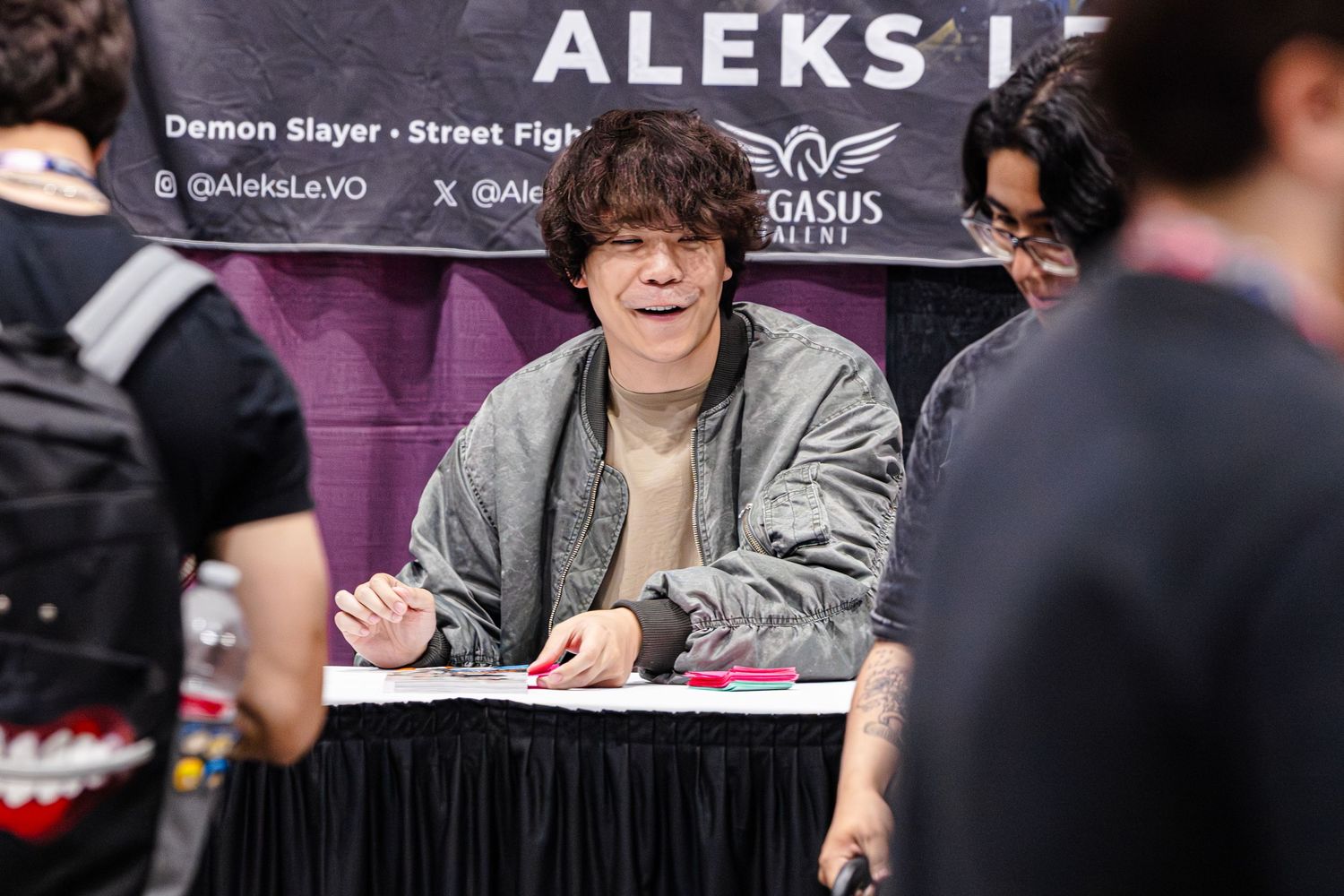 Person signing autographs at Aleks Le talent table at a gaming or anime convention event.