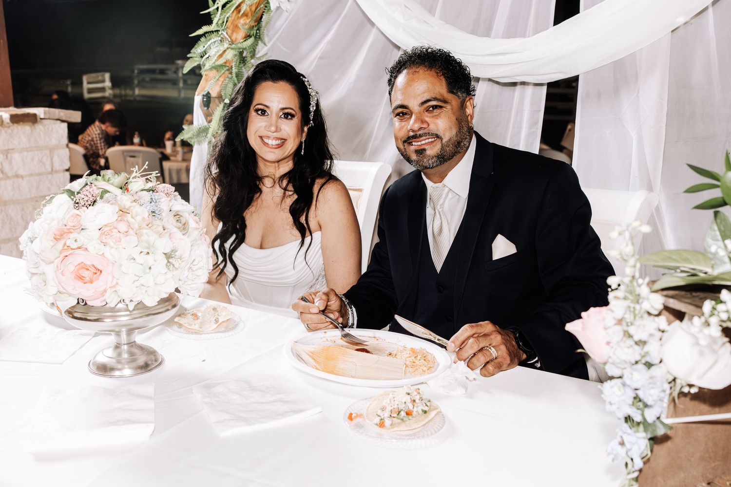 Wedding guests enjoy an elegant reception dinner at tables decorated with white linens and floral centerpieces.
