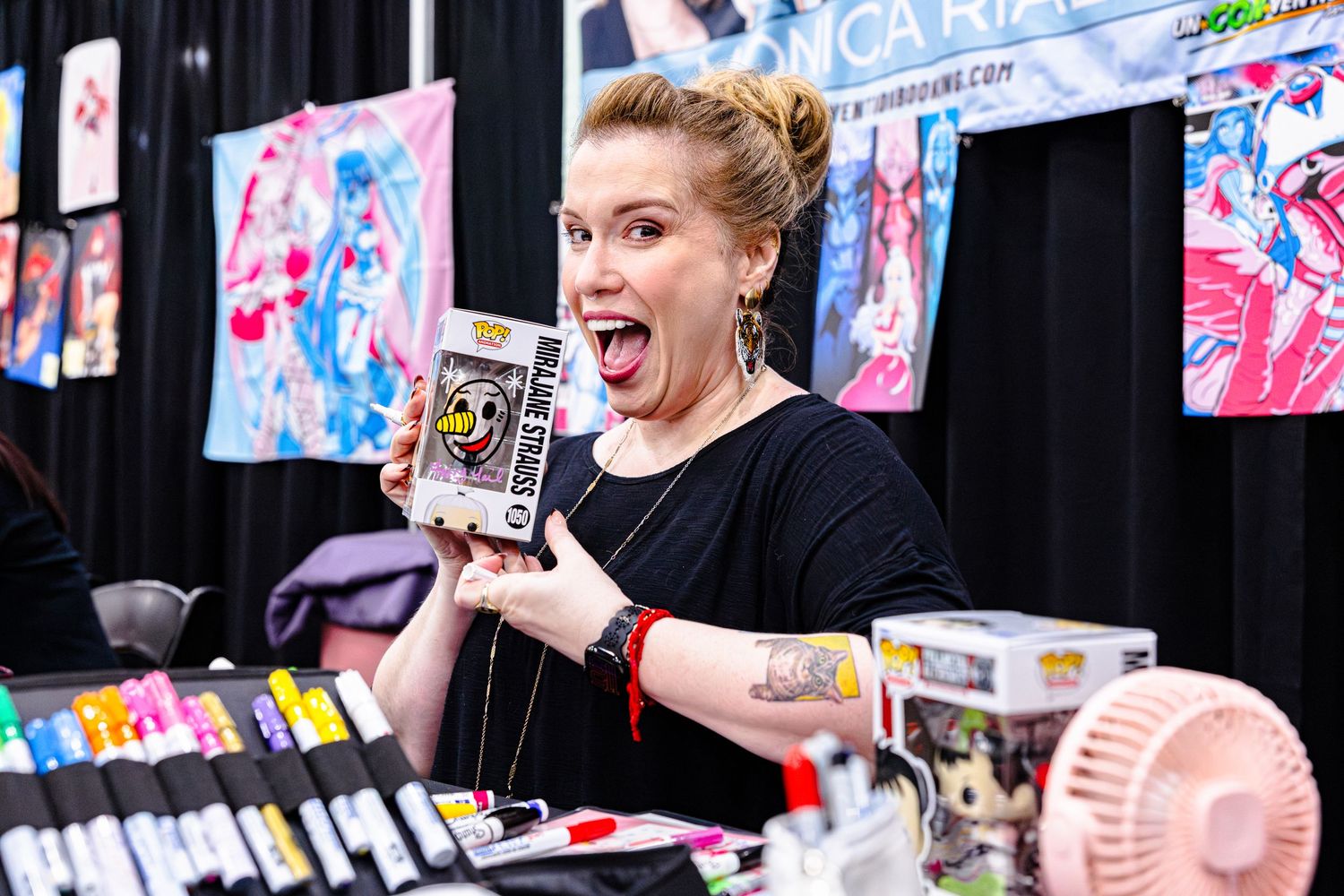 Excited woman holds Funko Pop box at anime art vendor booth surrounded by markers and collectibles.