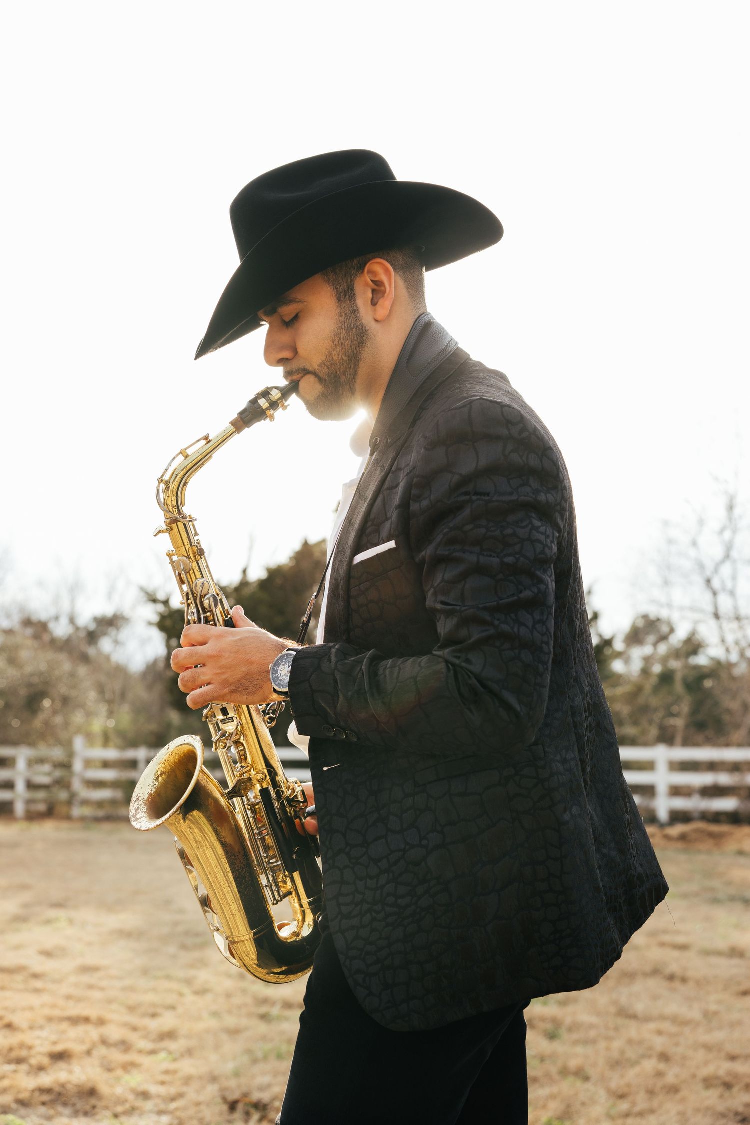 Musician in black suit and cowboy hat plays saxophone outdoors against rustic fence background.