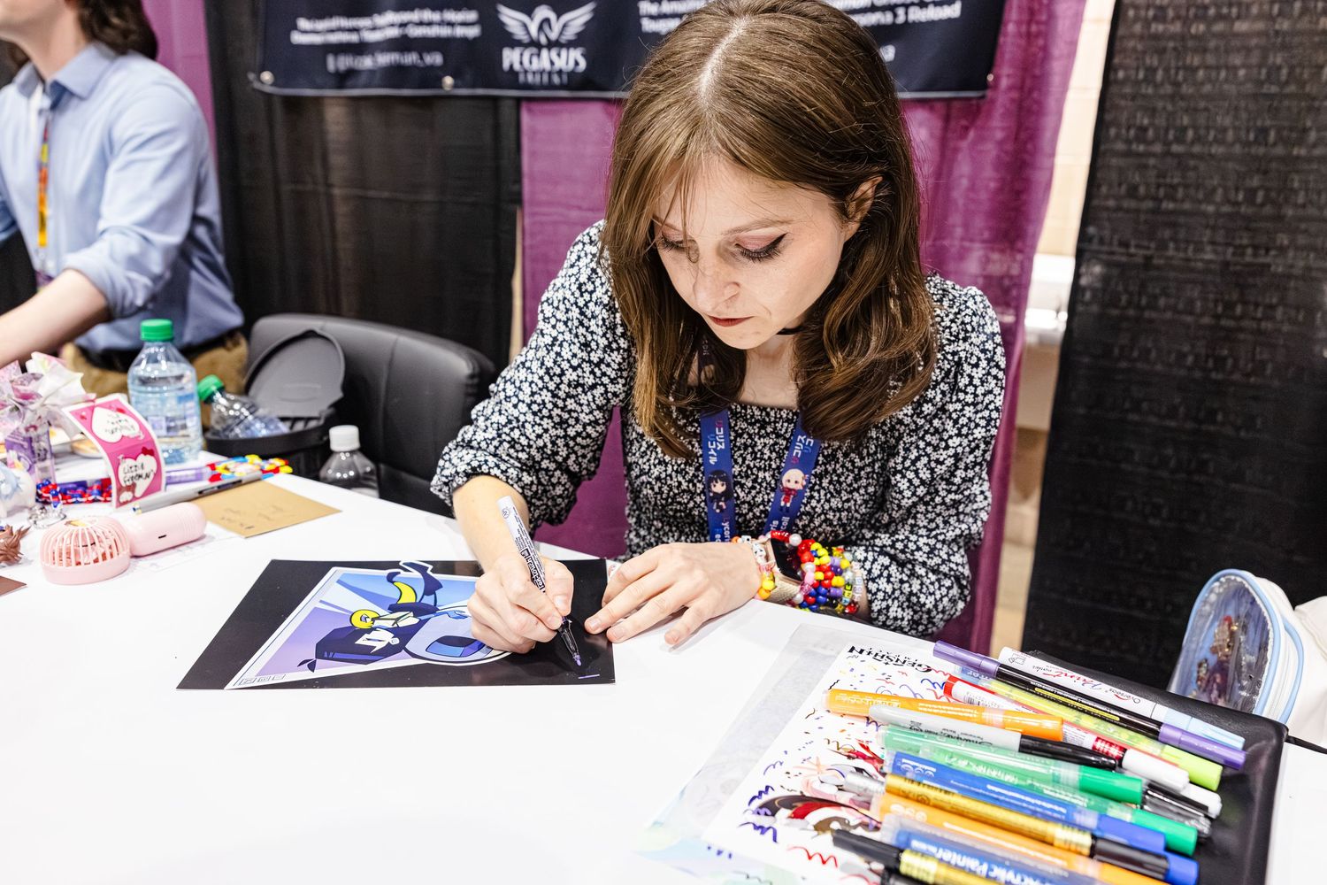 Young girl drawing colorful artwork at a convention booth with markers and art supplies on display.