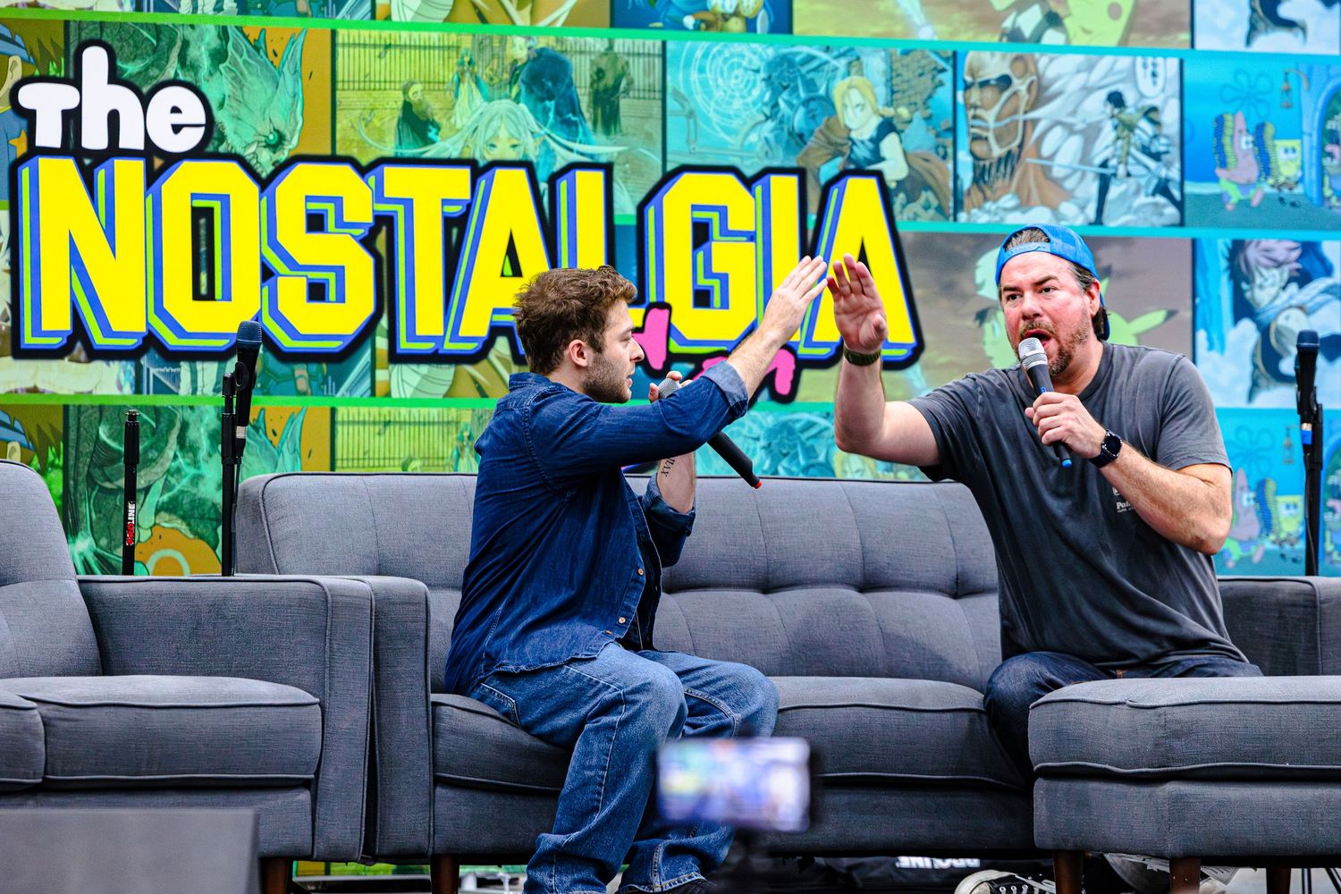 Two men high-five on stage at The Nostalgia event, seated on grey couches with colorful backdrop.