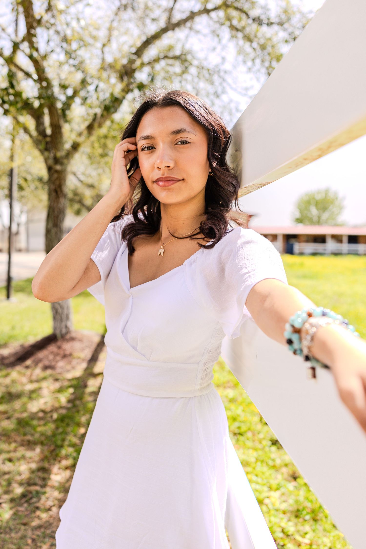 A person in a flowing white dress poses outdoors on a sunny day with trees in the background.