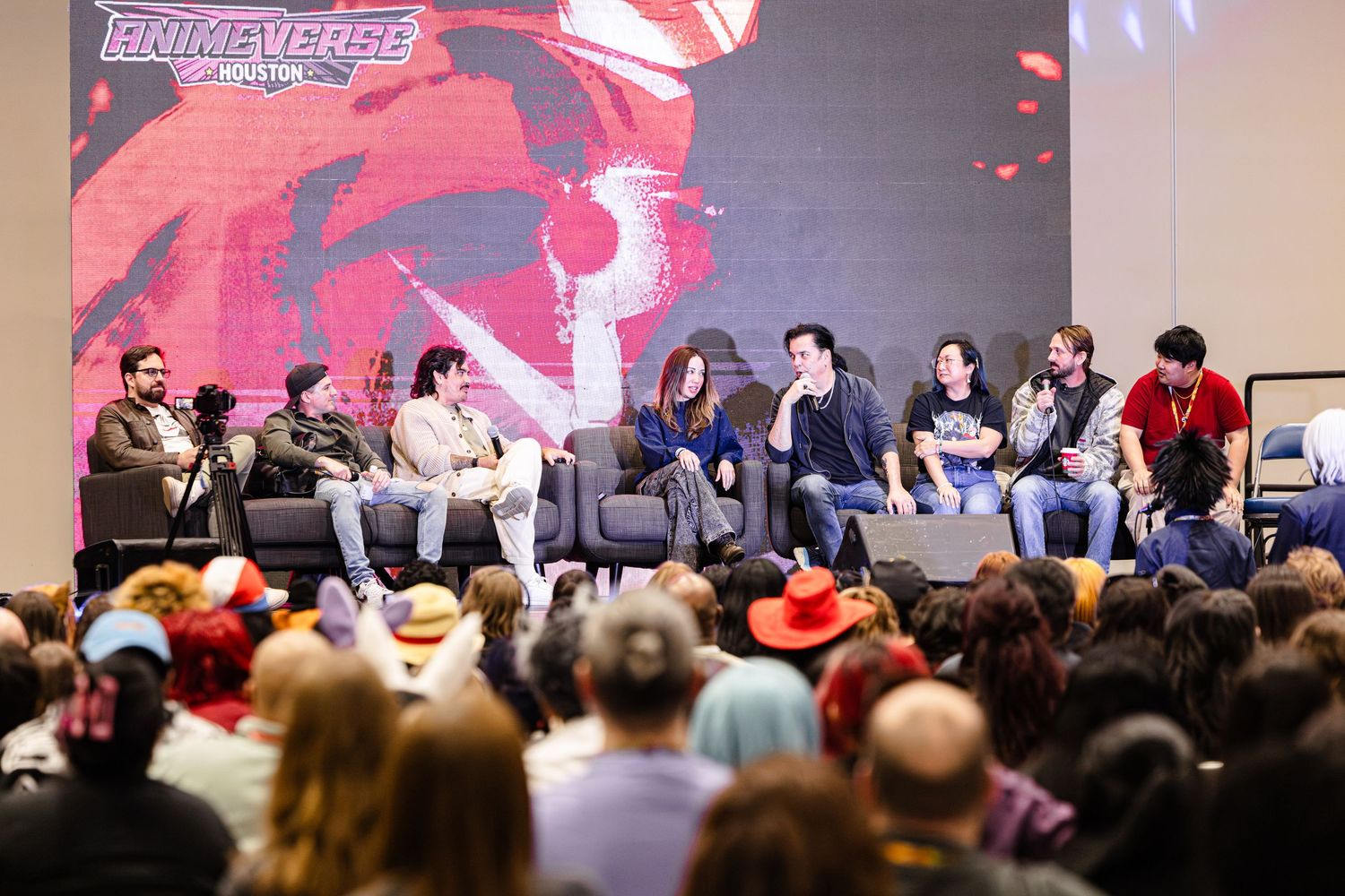 A panel of seven guests seated on stage at Primaverse Houston convention speaking to a large audience.