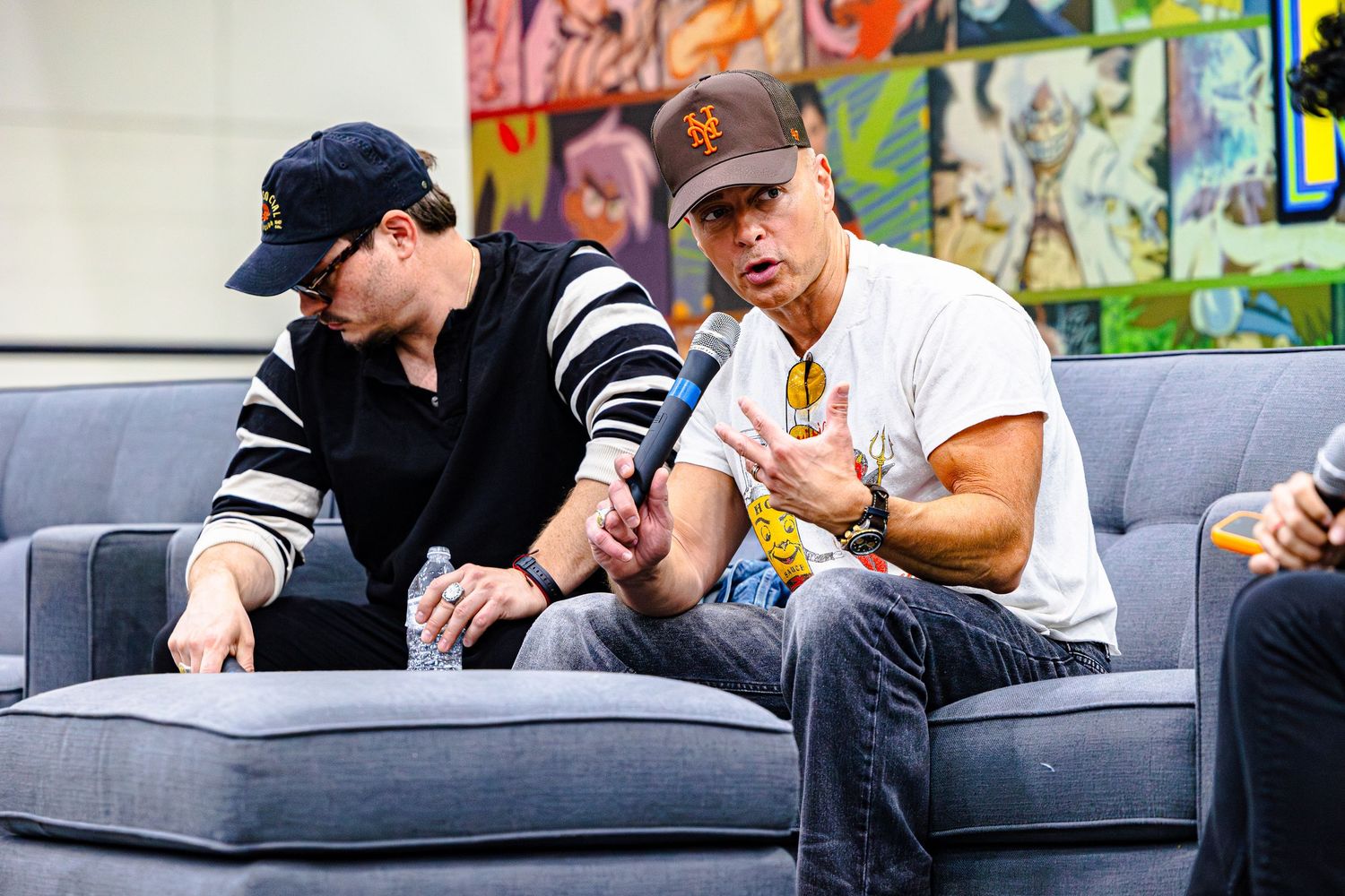 Two men sitting on a gray couch in conversation at what appears to be a comic convention panel event.