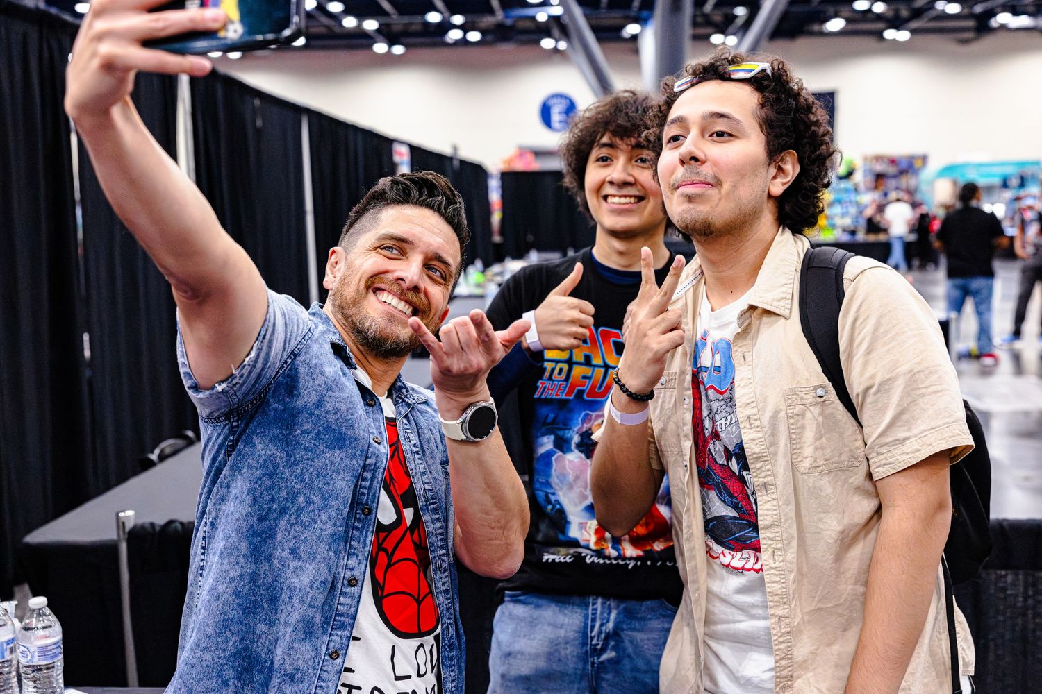 Three friends pose for a selfie at a comic convention, flashing peace signs and smiles indoors.