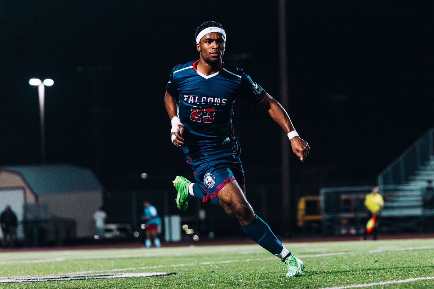 A soccer player in a navy blue Falcons jersey runs on a field at night under stadium lights.