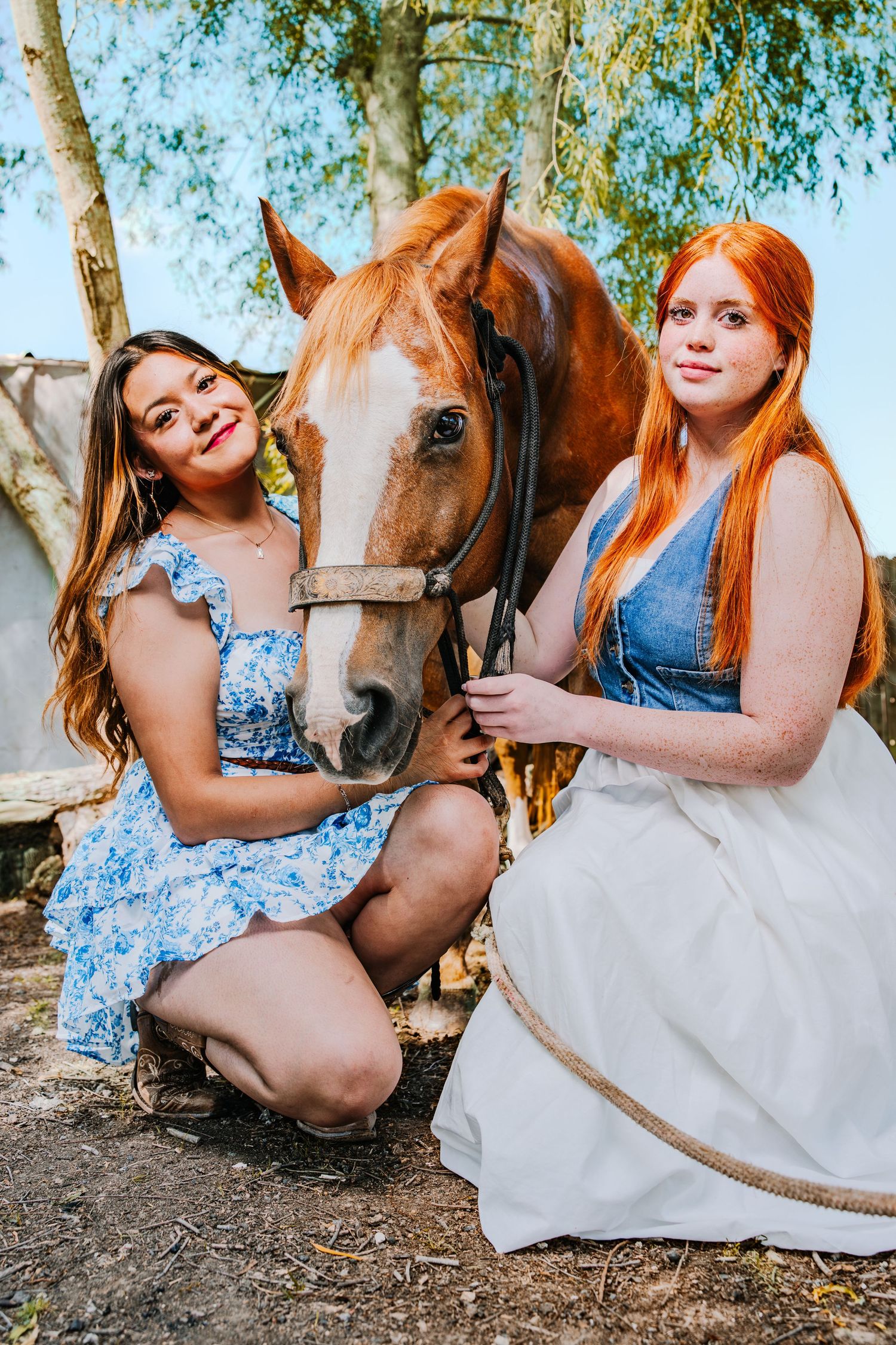 A brown horse and two people in blue and white dresses interact during a sunny outdoor photo session.