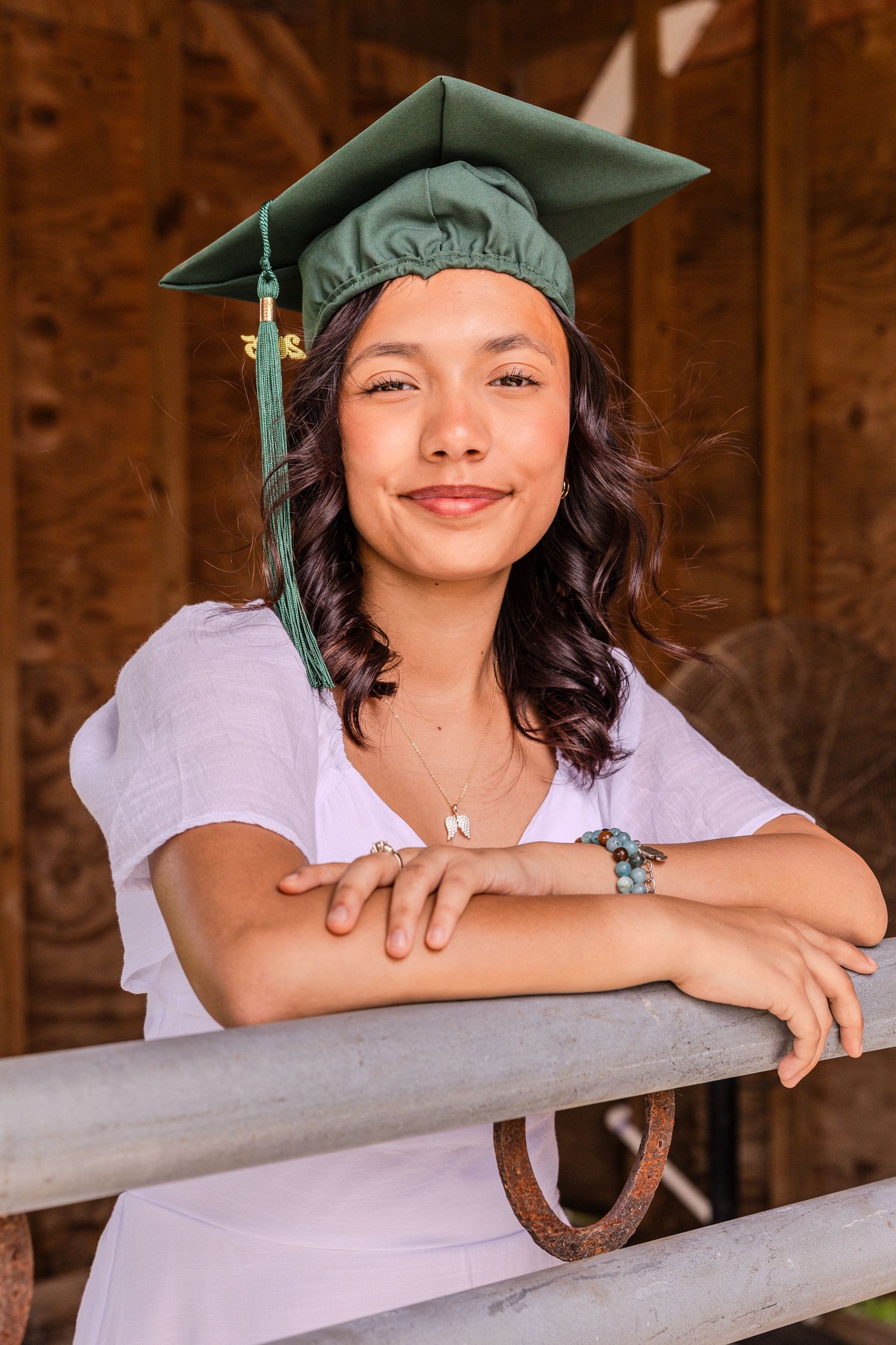 Graduate in green cap leans on wooden railing while posing for graduation photos in rustic barn setting.