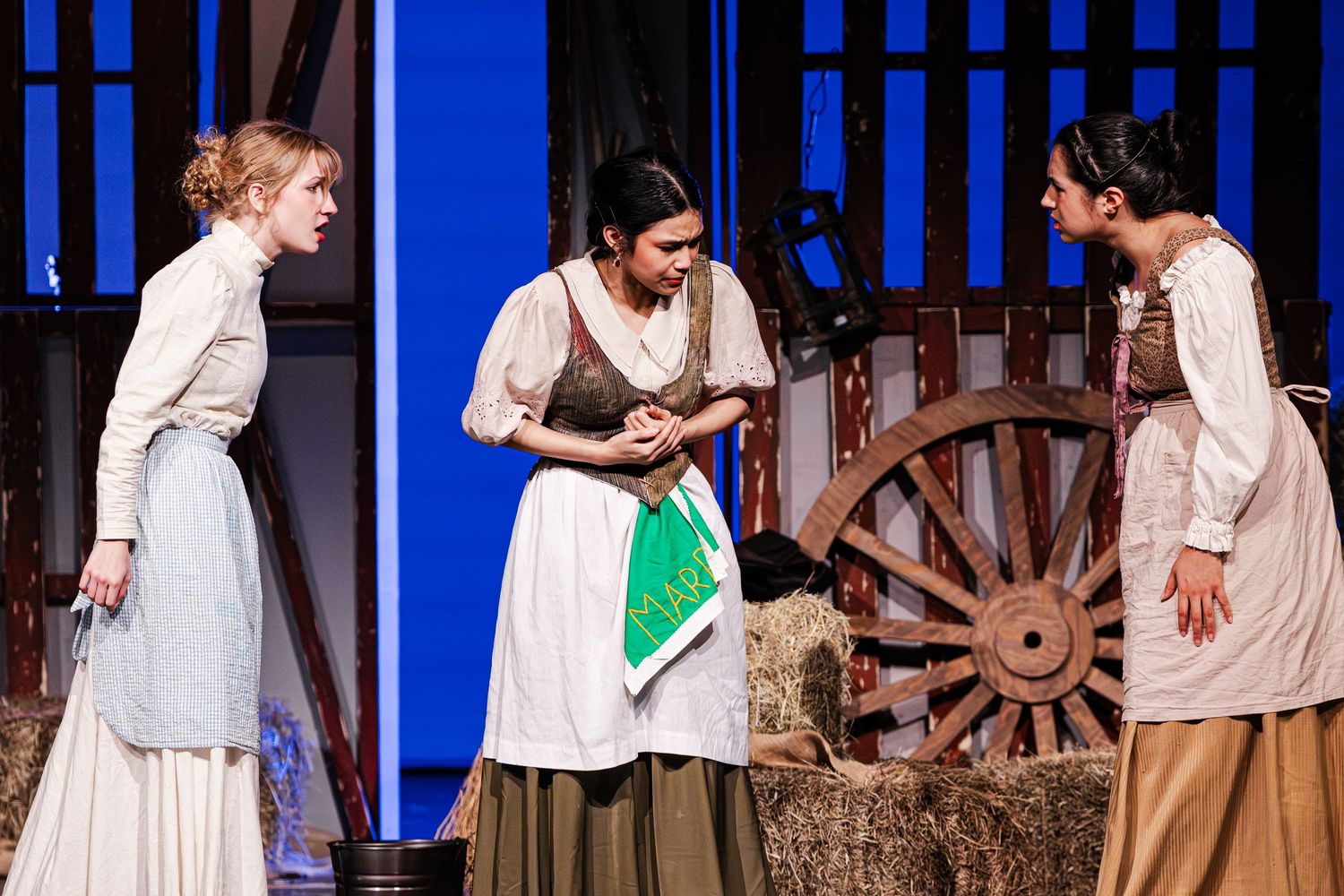 Three women in period costumes perform a theatrical scene on stage with hay bales and a wagon wheel backdrop.